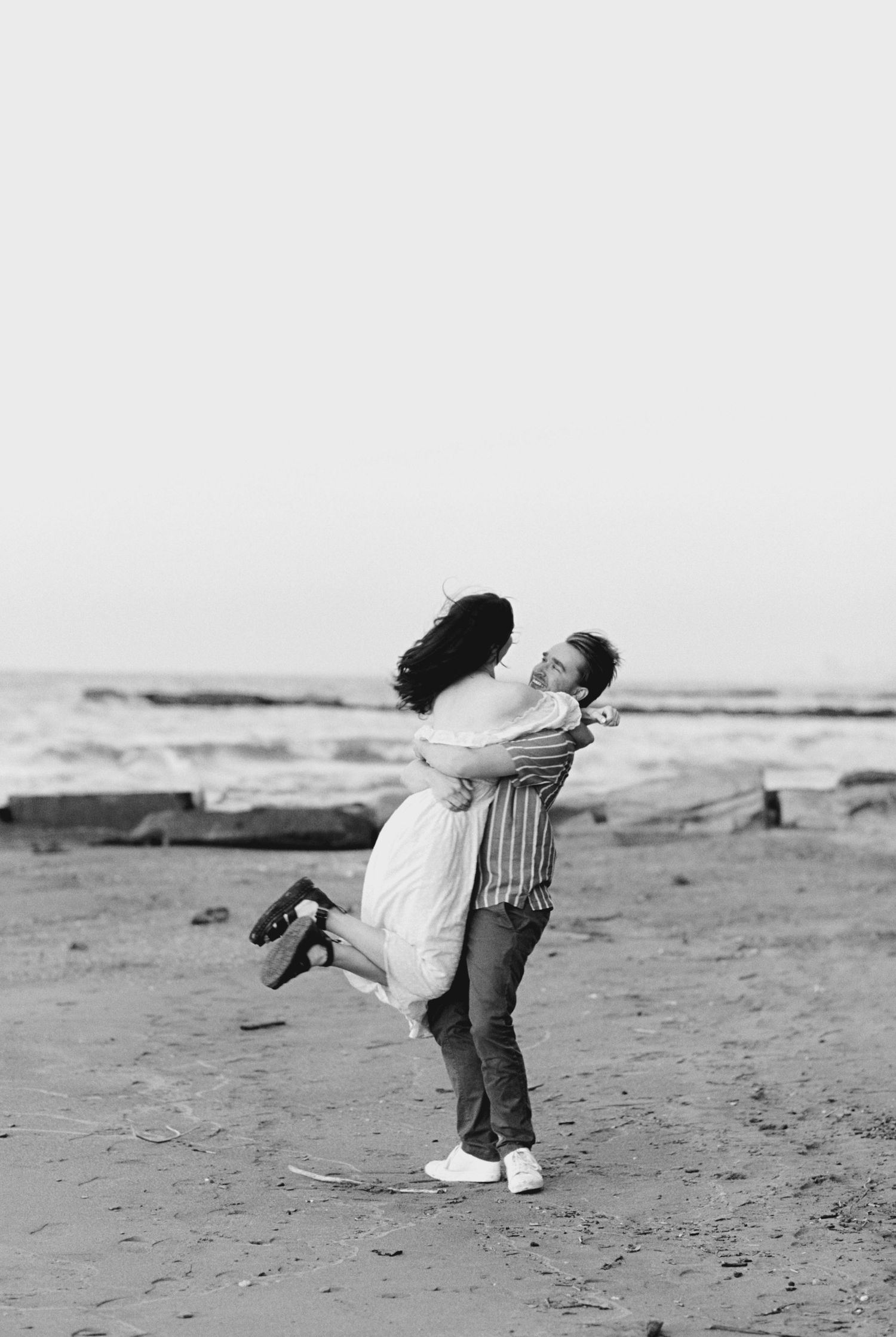 A couple shares a joyful embrace while standing on a beach, captured in black and white photography.