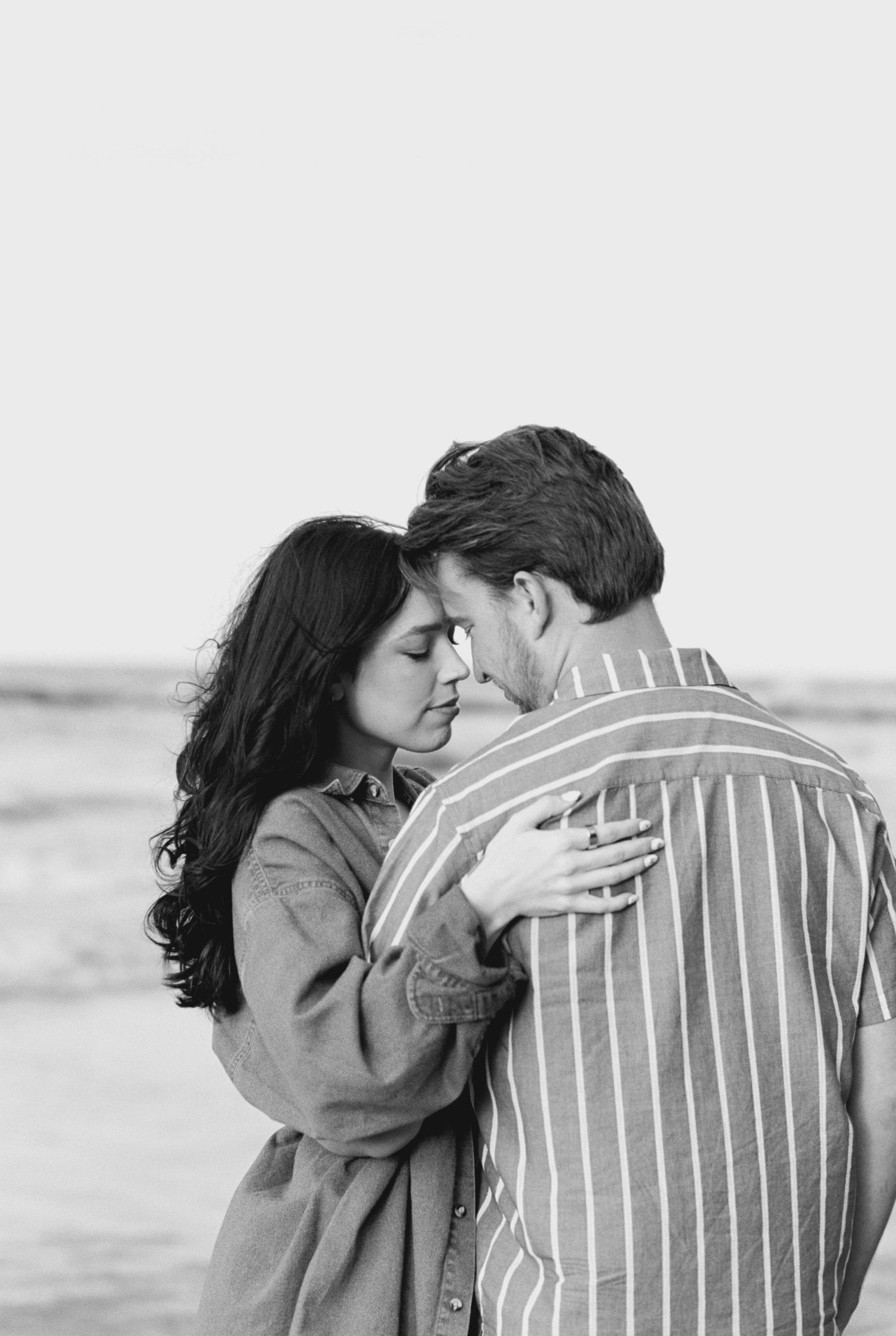A romantic black and white photo of a couple embracing on a beach, with one wearing a striped shirt and the other in a coat.