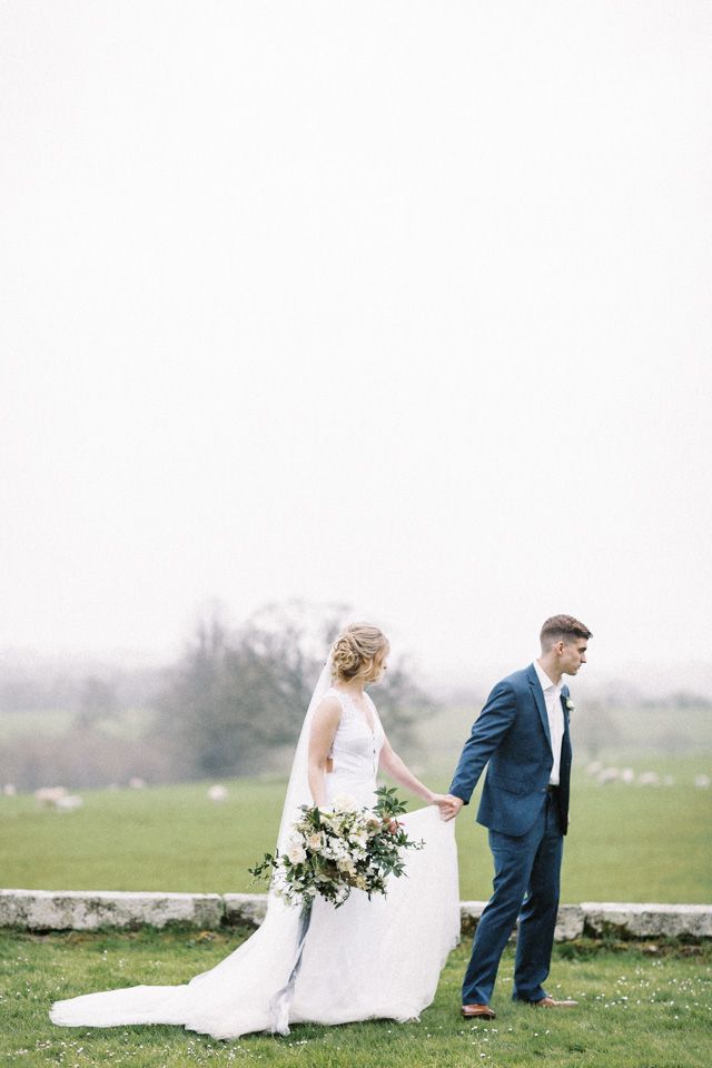 A couple stands outdoors in wedding attire with the bride holding a lush greenery bouquet against a misty countryside backdrop.