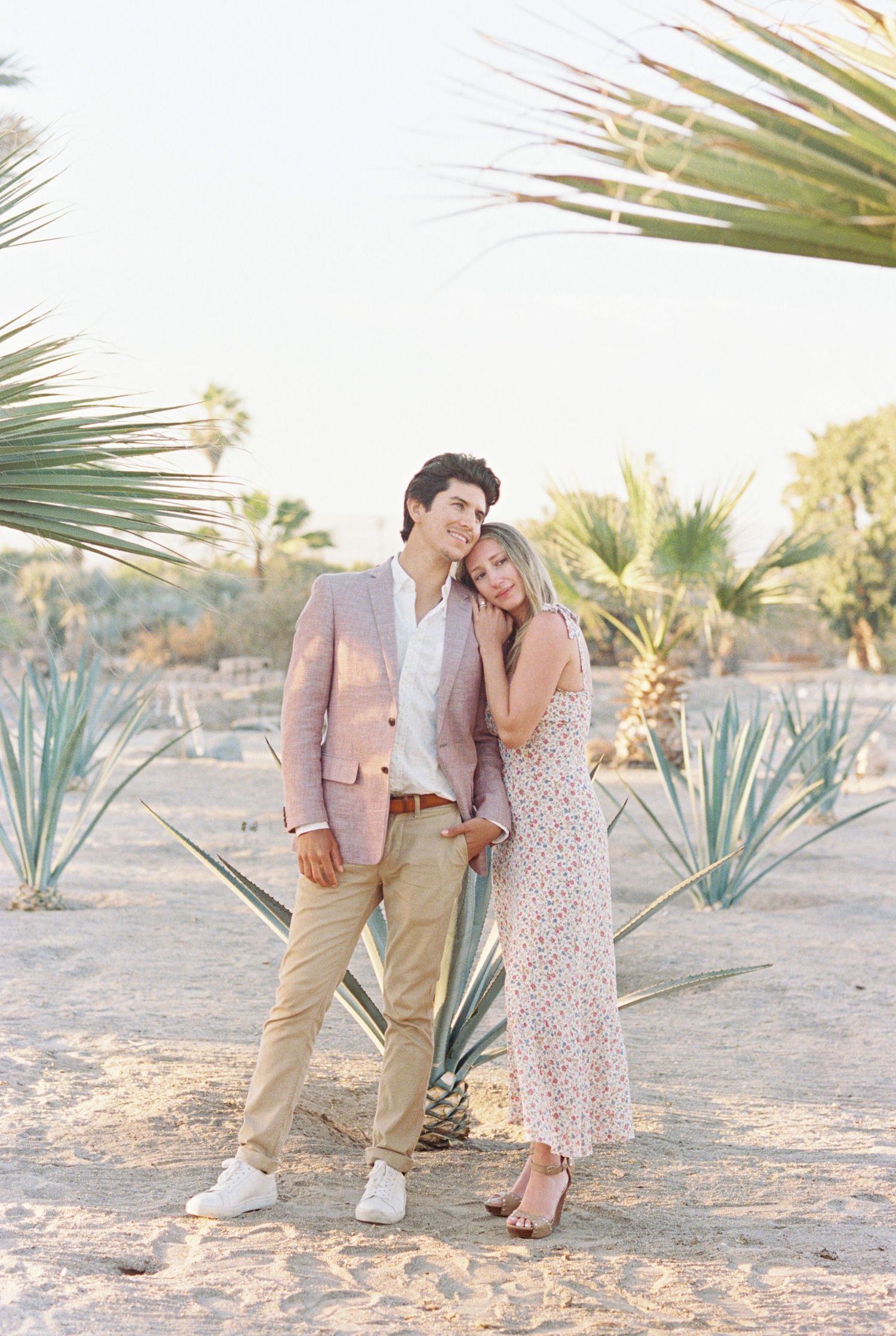 Romantic couple poses in desert landscape with agave plants and palm fronds, wearing pastel and neutral toned outfits. Mexico Wedding
