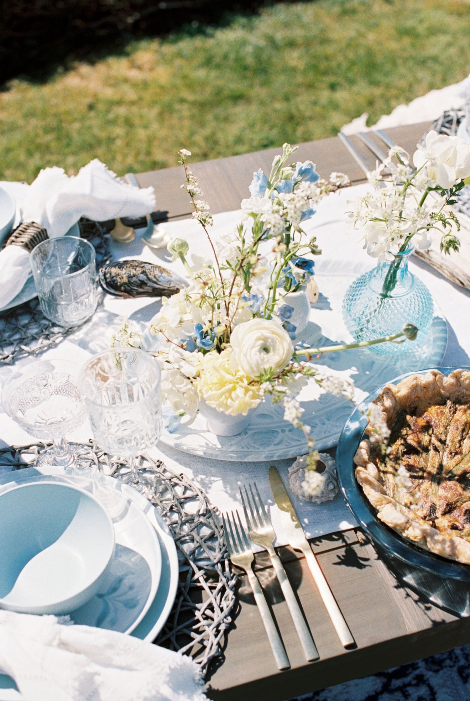 Elegant outdoor table setting with light blue plates, white linens, floral centerpieces and rustic accents in natural light.
