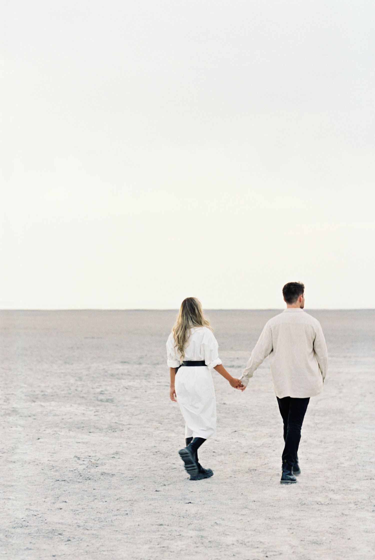 A couple holding hands while walking on an empty salt flat desert in white and neutral colored outfits.
