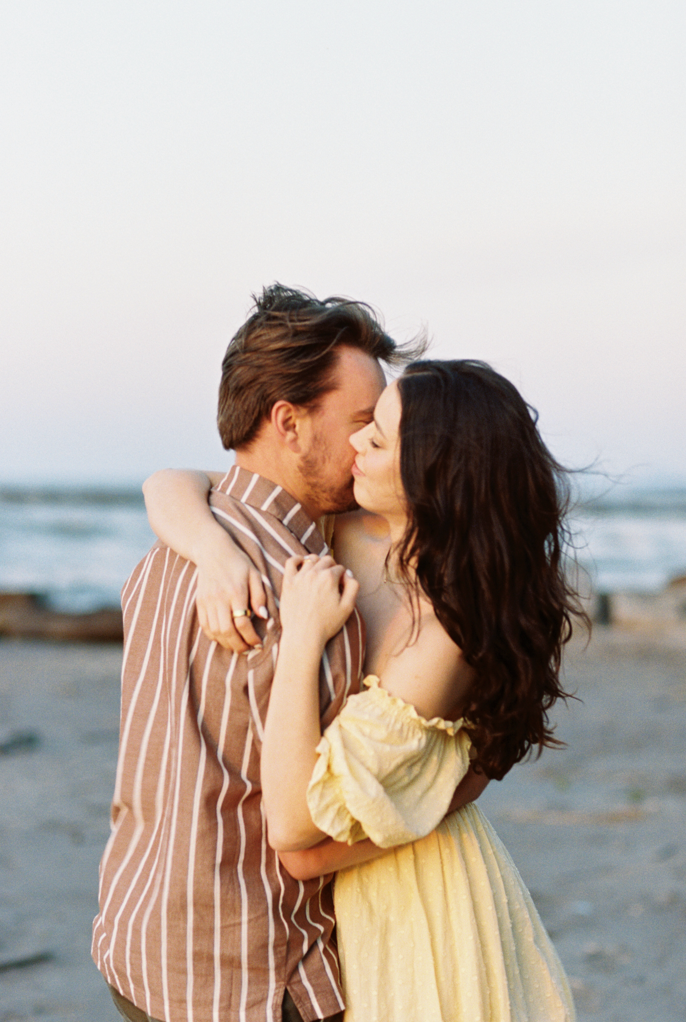 A romantic couple embraces and kisses on a beach at sunset with the ocean waves visible in the background.