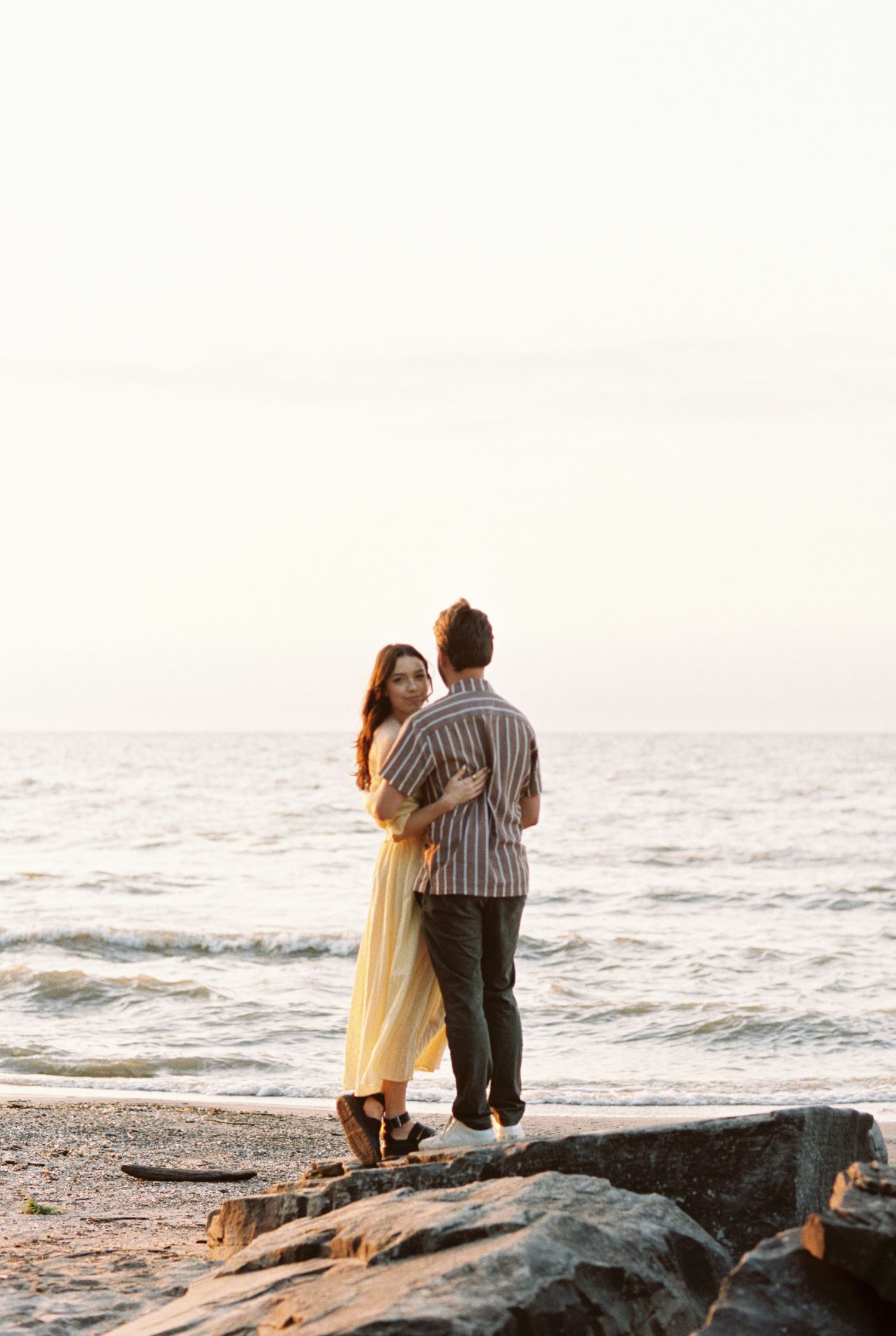 Two people stand together on rocky coastal outcrop at sunset overlooking the ocean waves.