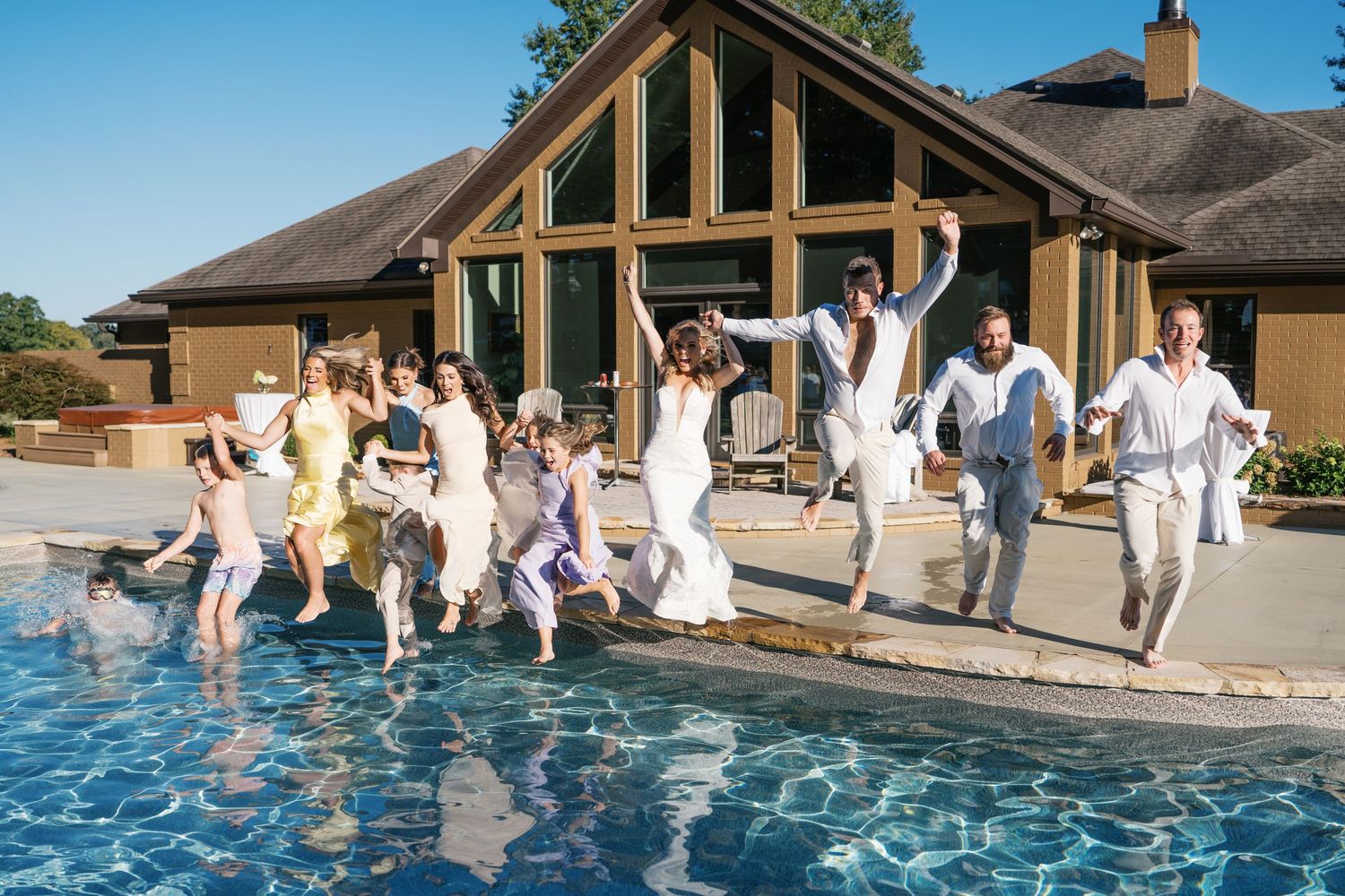 A group of people jump into a swimming pool in a synchronized line formation on a sunny day at a resort.
