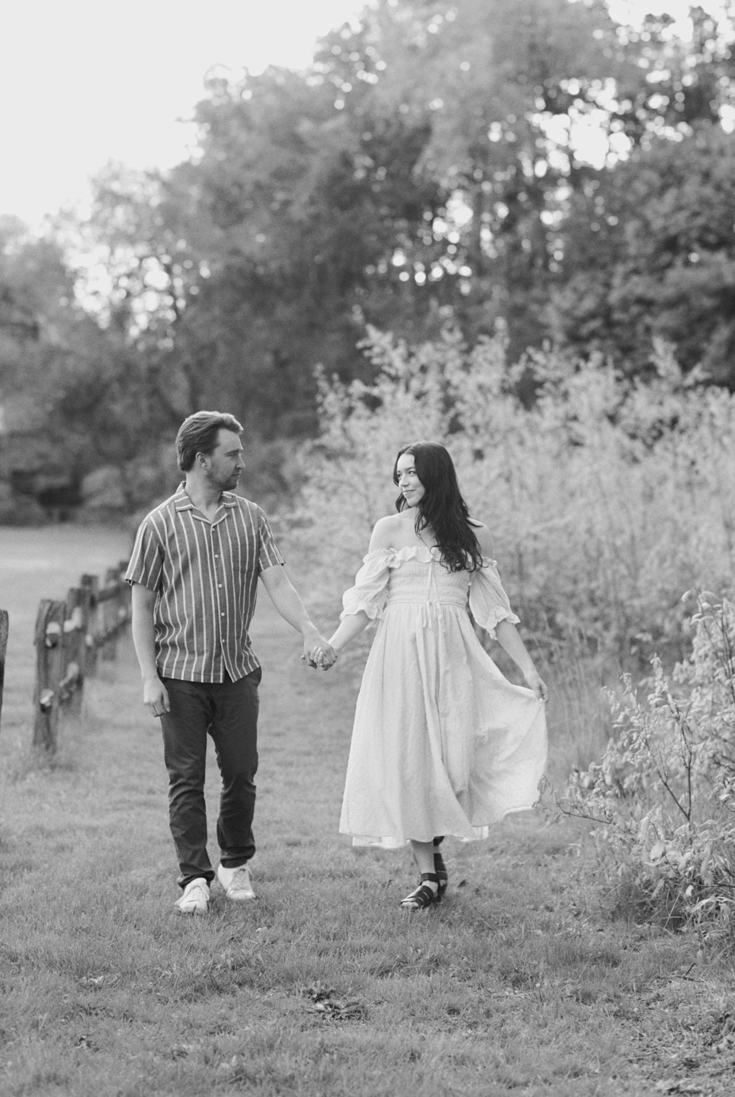 A romantic black and white countryside photo of a couple walking hand in hand on a grassy path near a rustic fence.