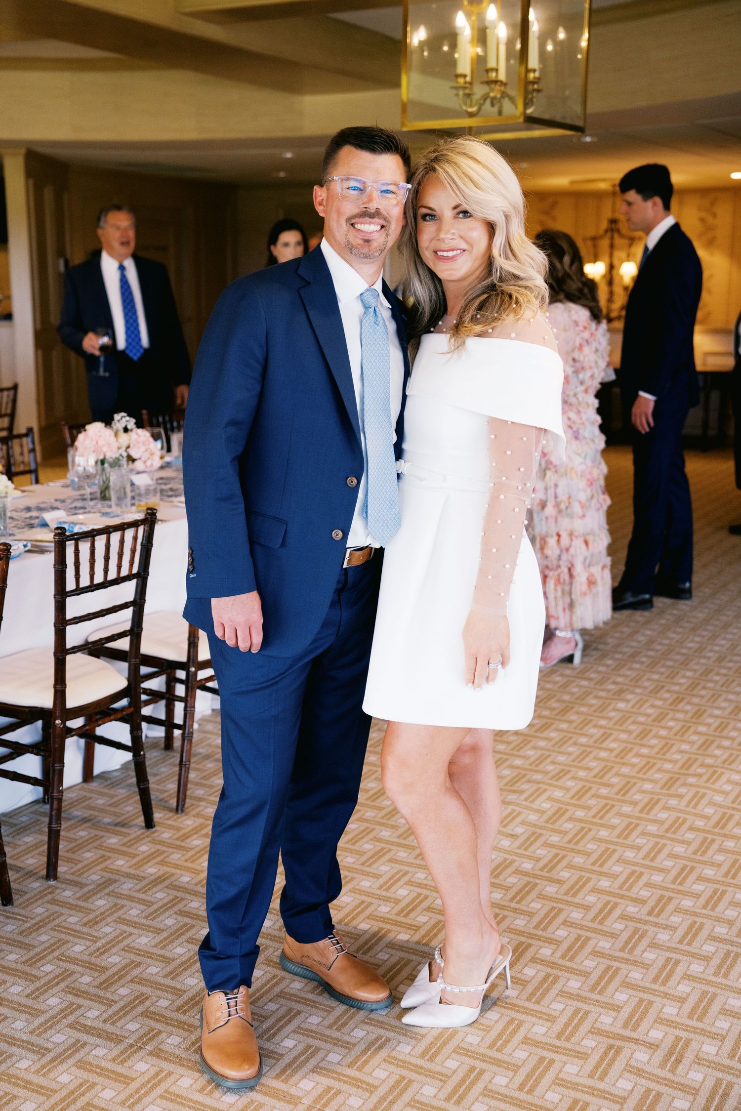 A couple poses at an elegant event, with the man in a navy suit and the woman wearing a white short dress.