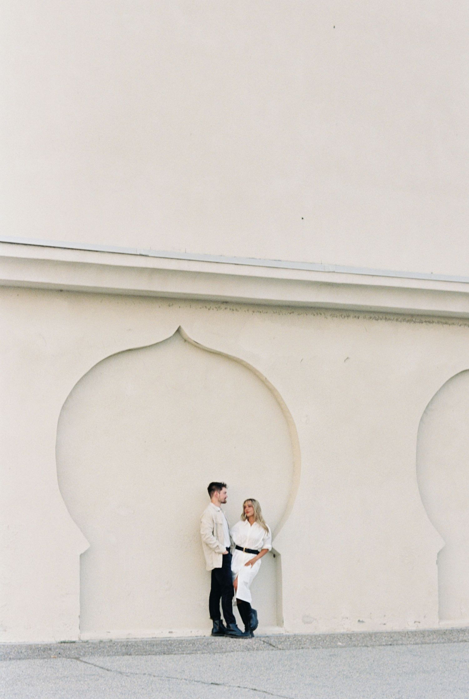 A couple stands together inside an arched alcove with Moorish architectural details against a cream-colored wall.