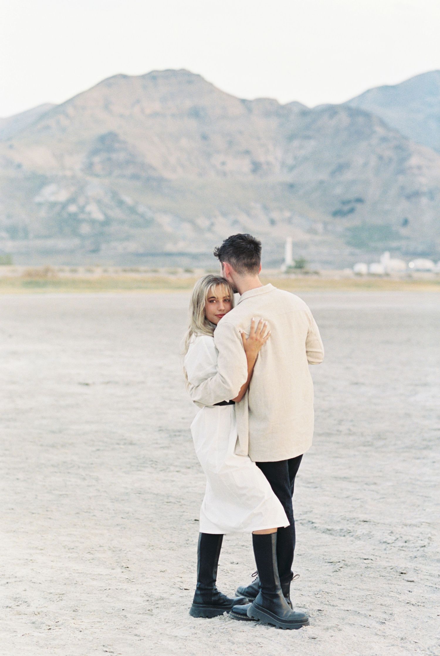 A couple embraces on a desert plain with mountains in the background on an overcast day.