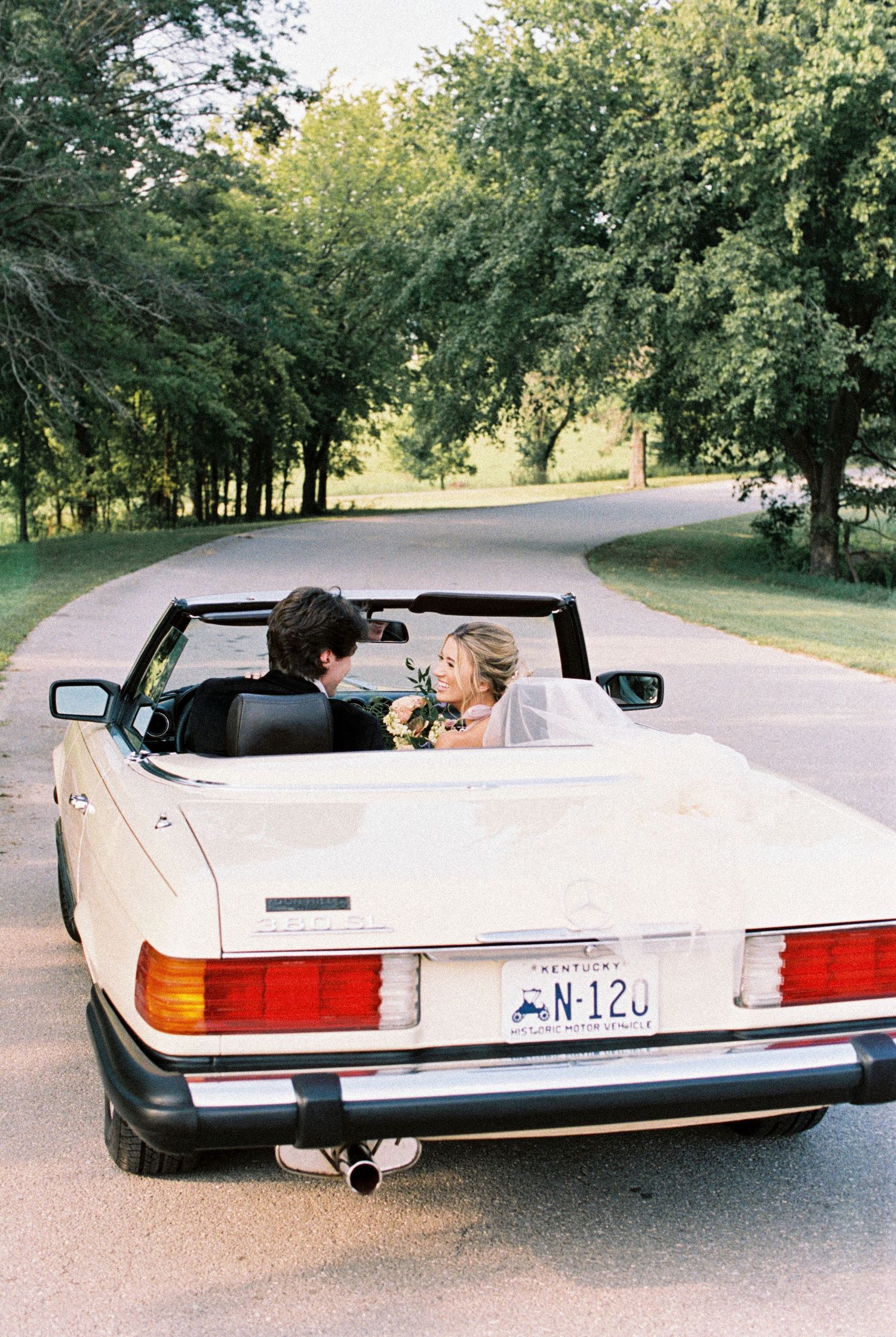 A white classic Mercedes convertible drives down a tree-lined road on a sunny summer day.