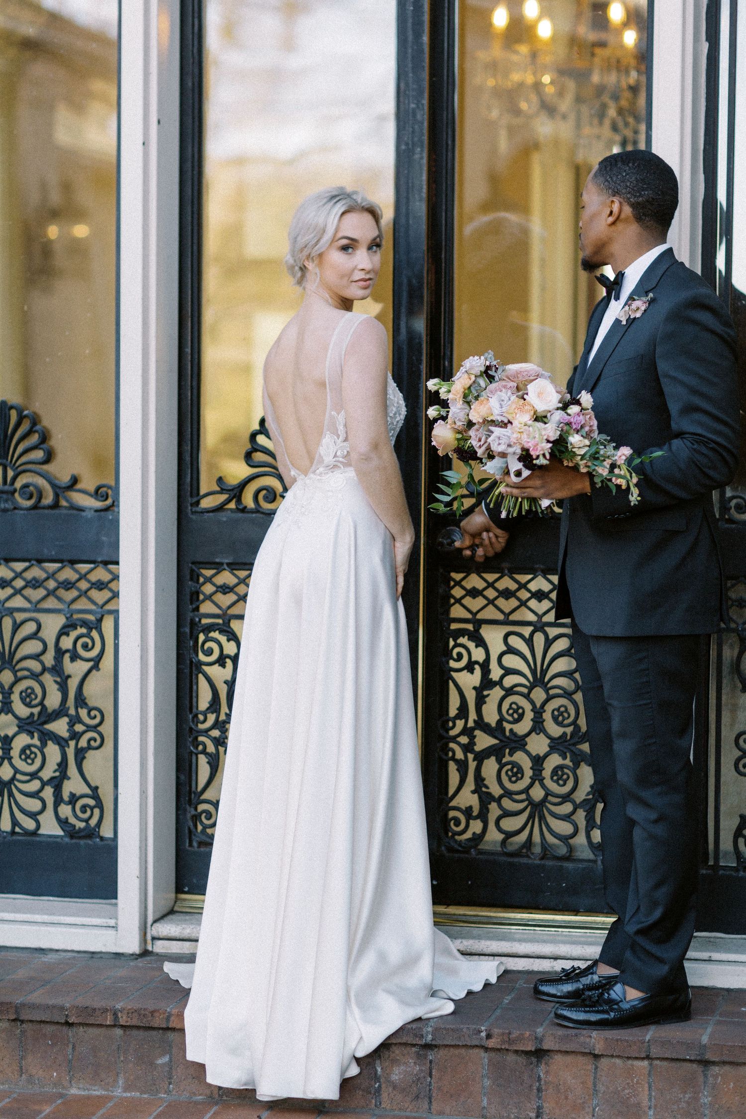 A romantic wedding photo with a couple in formal attire posing by ornate glass doors with pink floral bouquet. Kirstie Whitaker Photography