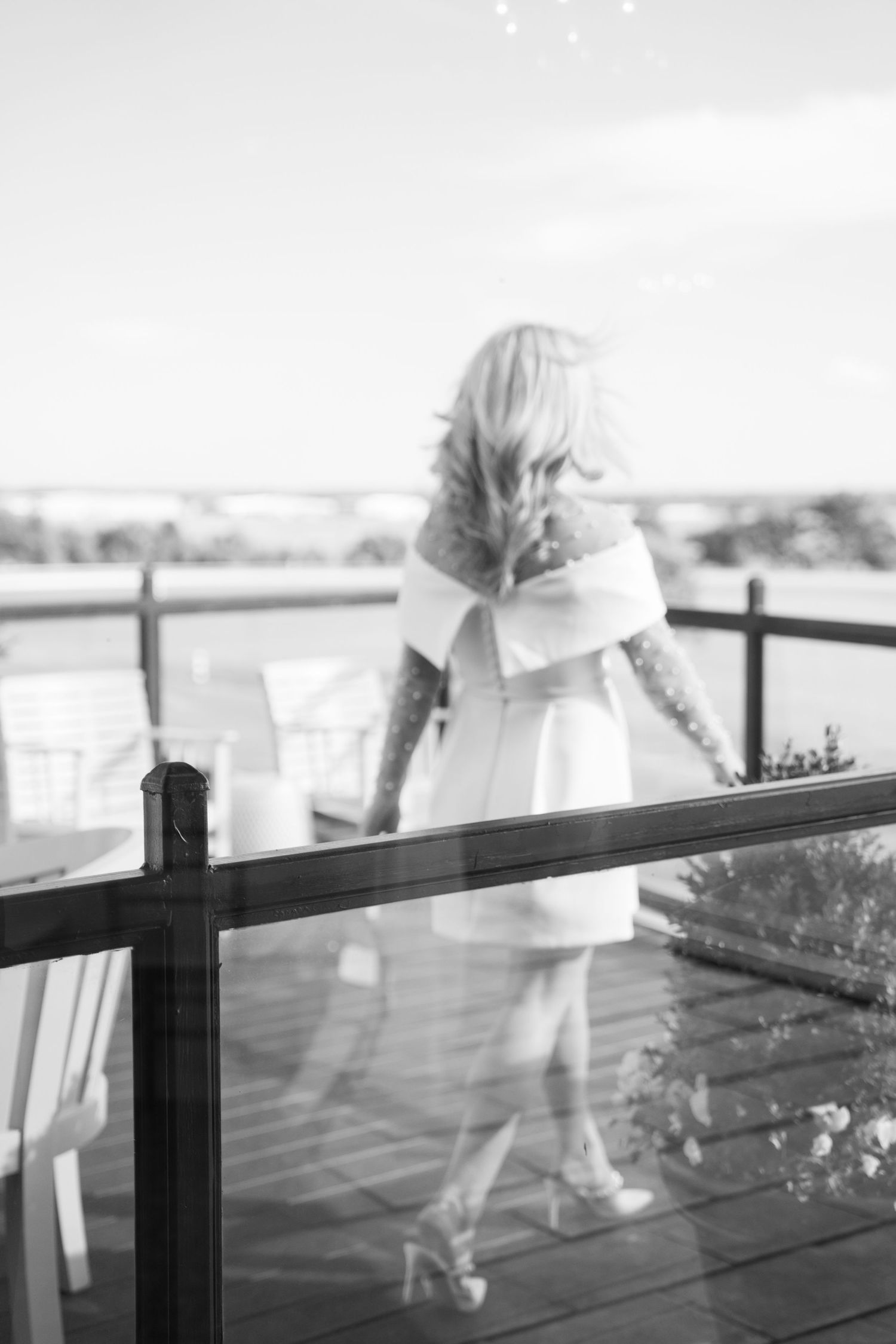 Black and white photo of figure in white dress standing on wooden deck overlooking scenic outdoor vista.