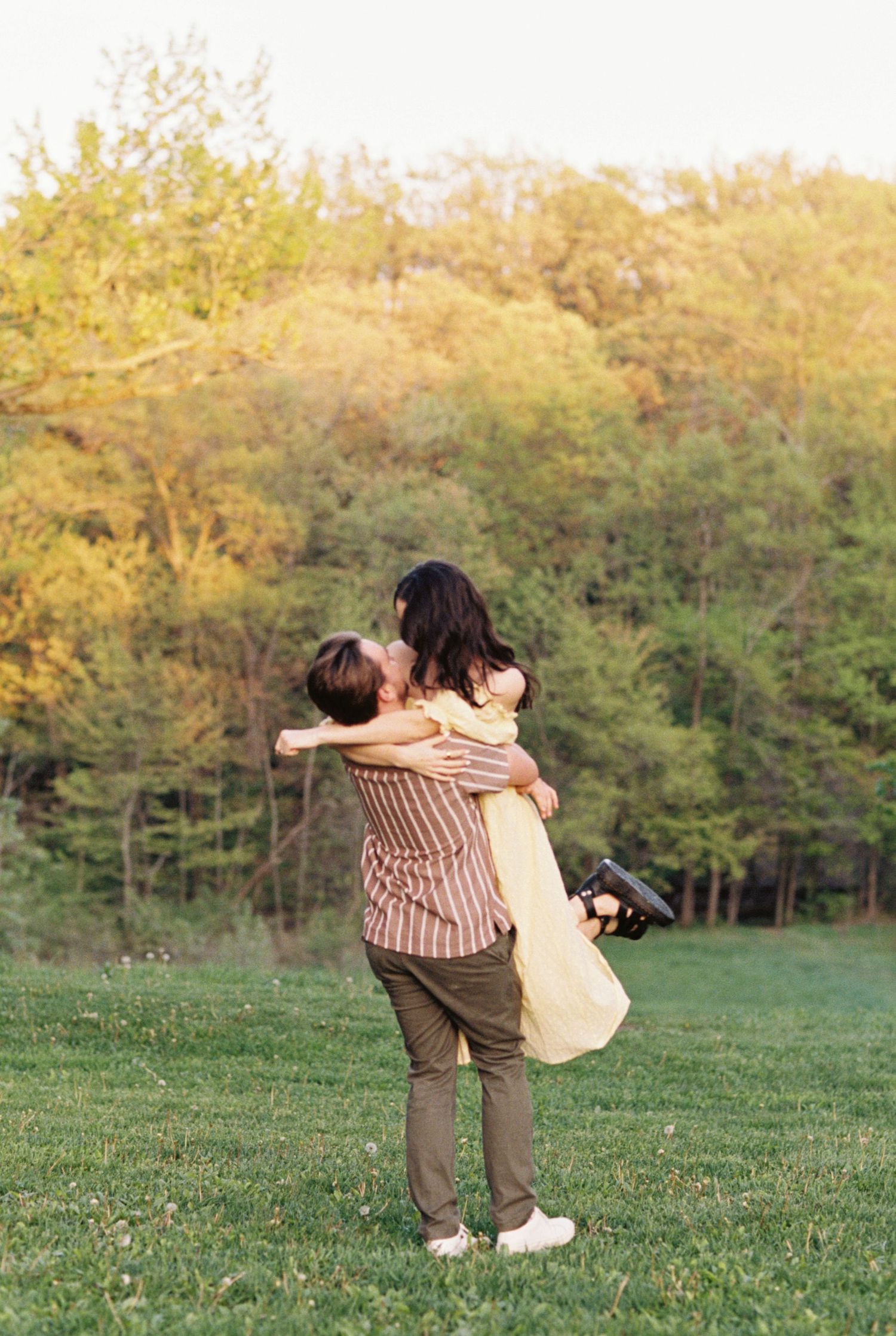 A couple embraces in a tender moment on a grassy hillside during golden hour with trees in the background.
