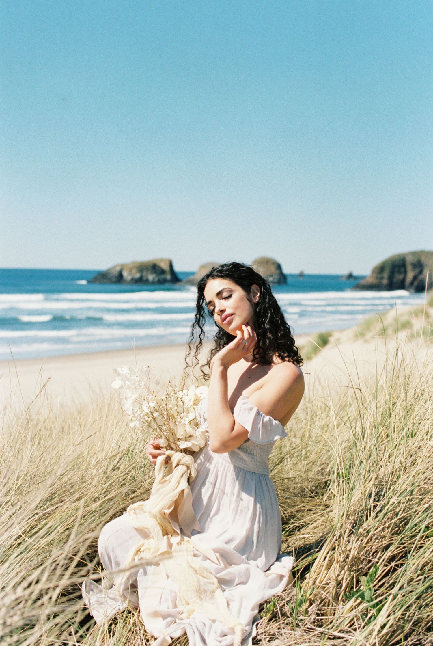 White summer dress flows in coastal breeze as figure sits among beach grass with ocean and rock formations in background.