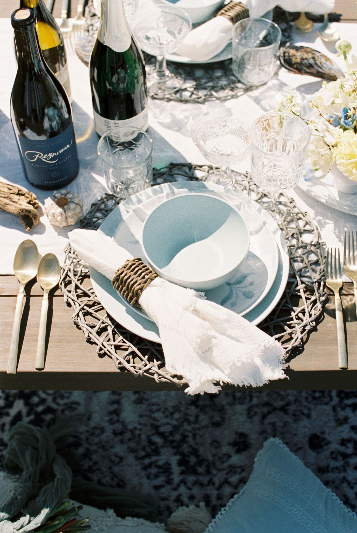 Elegant table setting with blue plates, wine bottles, and silver charger plates decorated with lace napkins and gold cutlery.