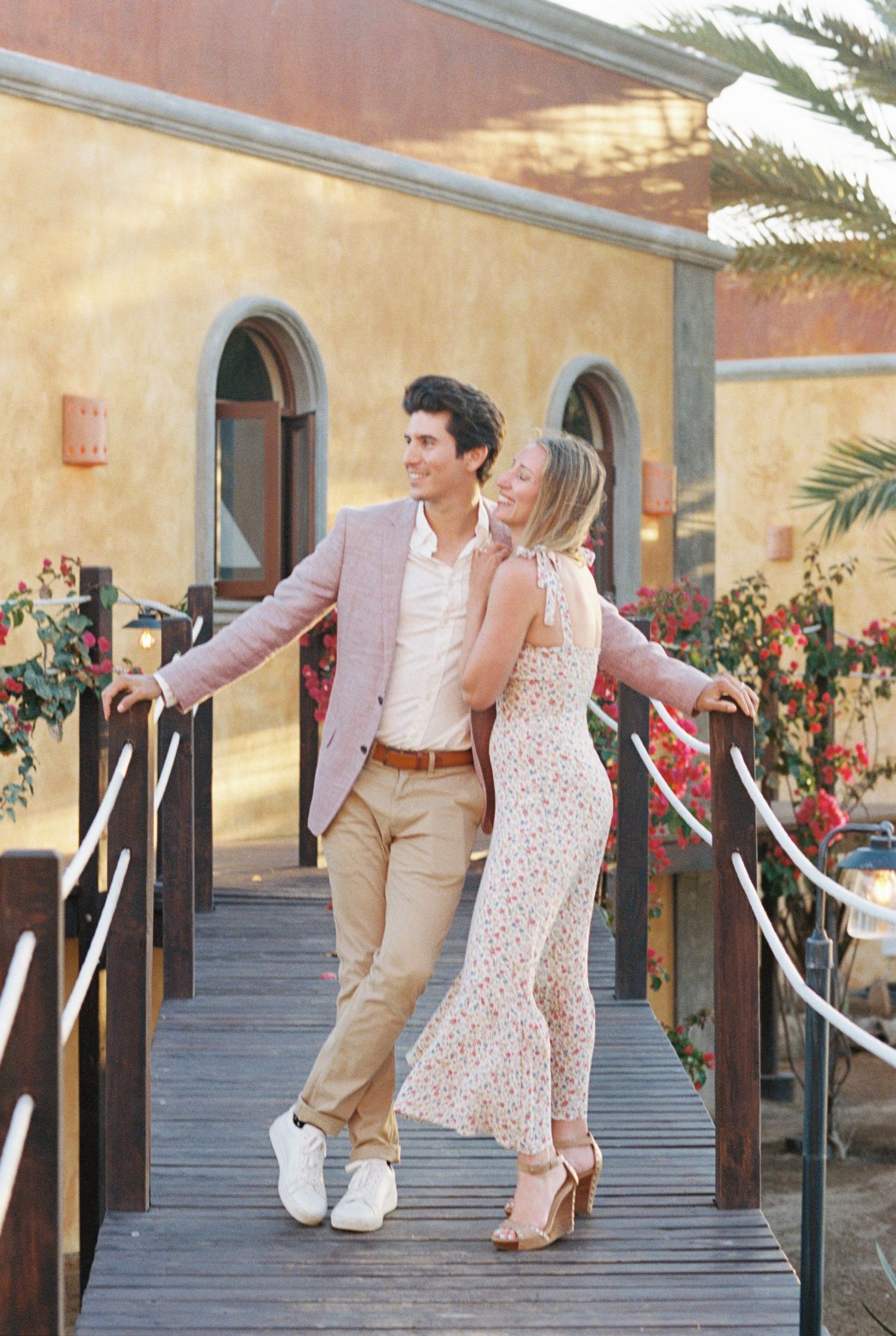 A couple in elegant attire pose together on a wooden bridge walkway against a Mediterranean-style building with palm trees.