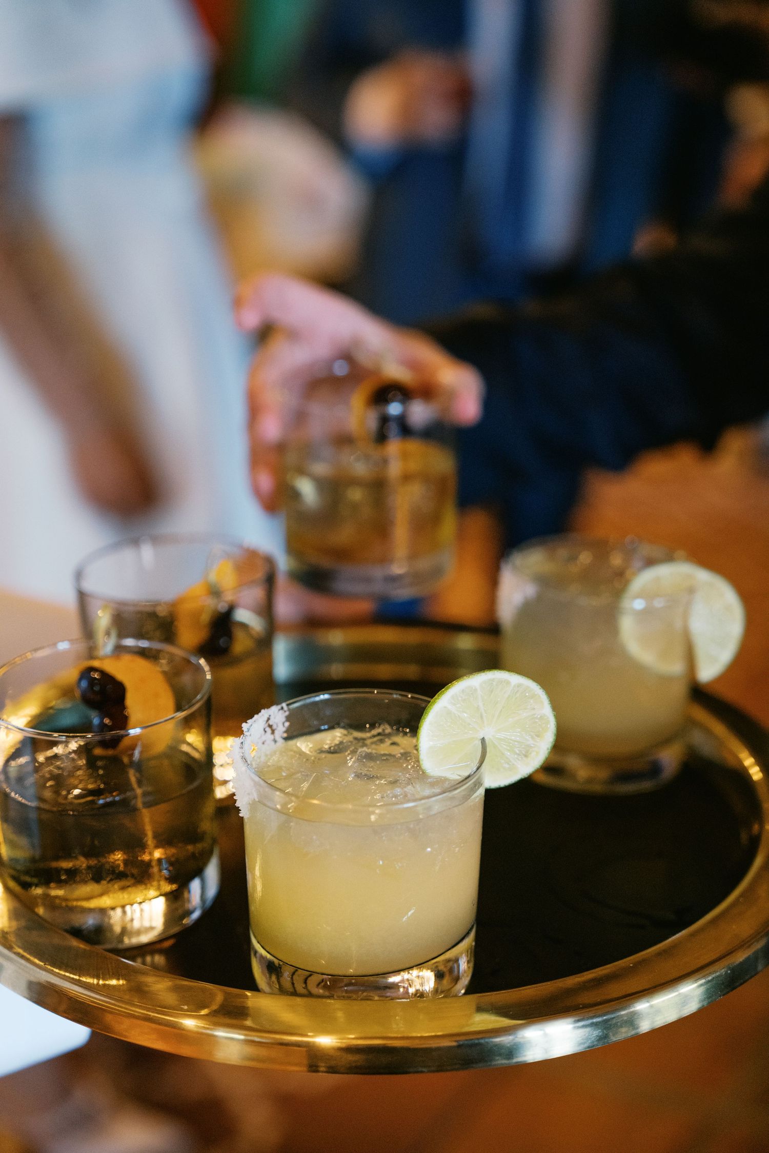 A round gold serving tray holds elegant cocktails garnished with lime wedges and olives in a dimly lit bar setting.