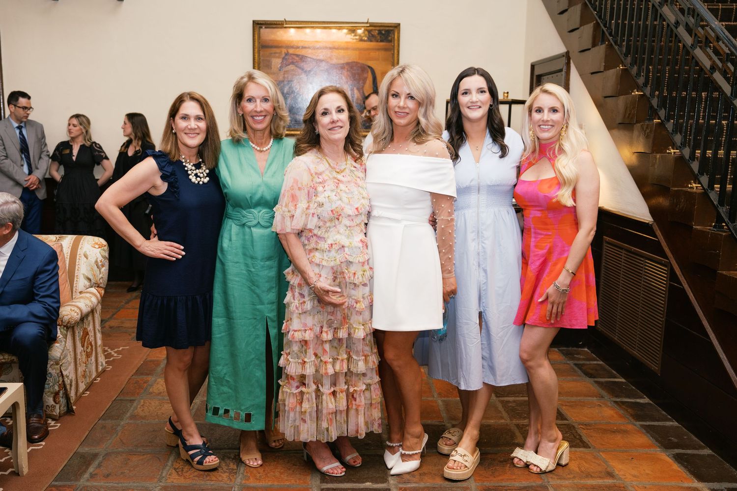 A group of women in stylish summer dresses gather together for a photo at a formal event with a decorative staircase visible.