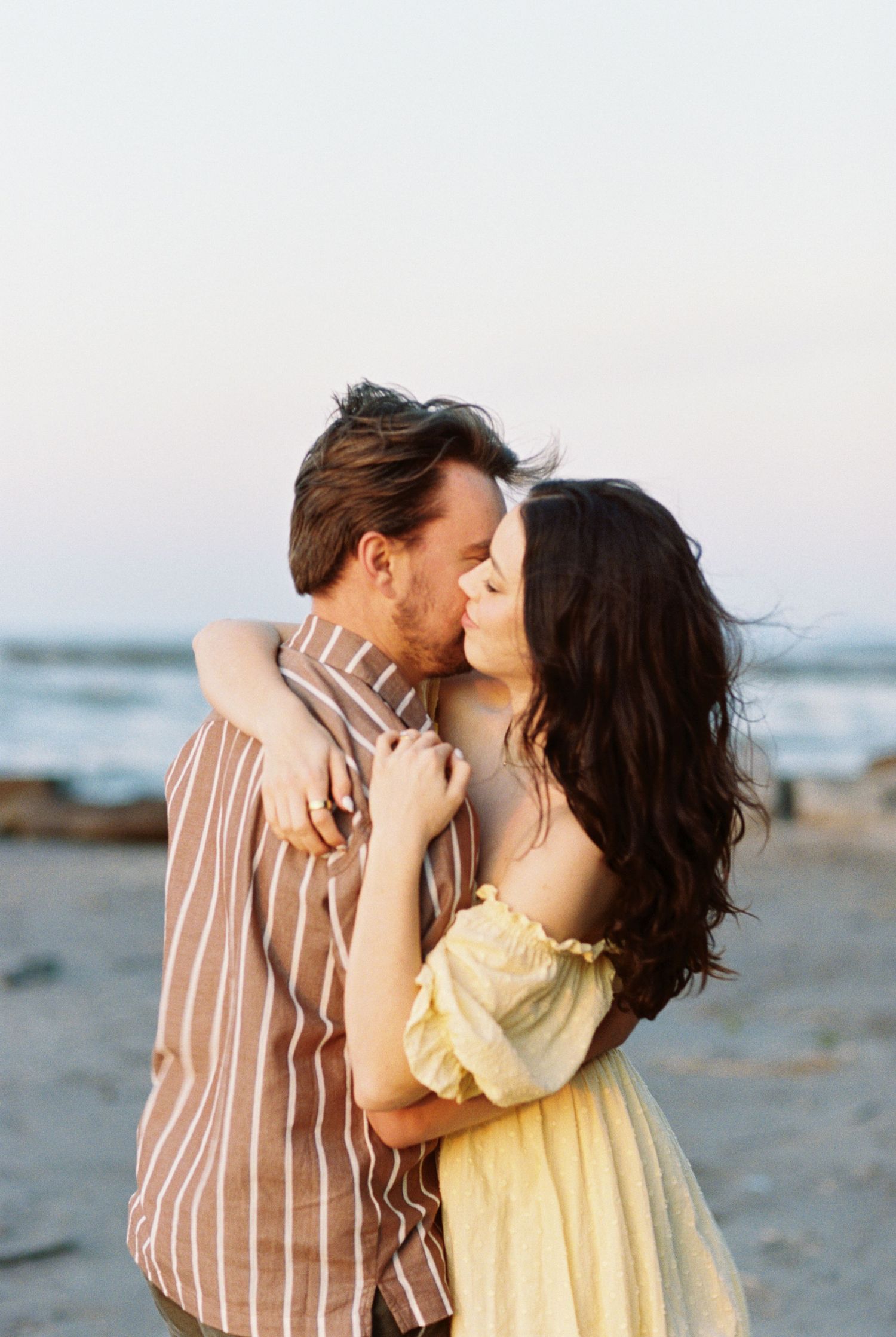 A romantic couple embraces on a beach at sunset, with her in a yellow dress and him in a striped shirt.