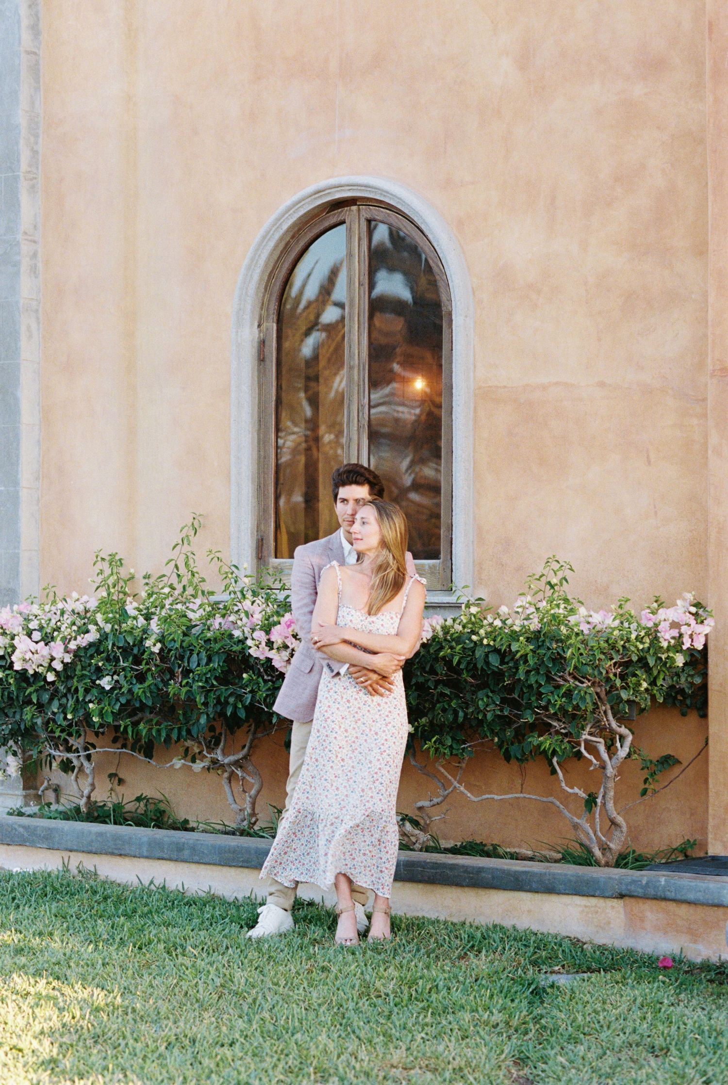 Couple embracing by arched window with flowering window box in Mediterranean setting, Mexico Wedding Photographer