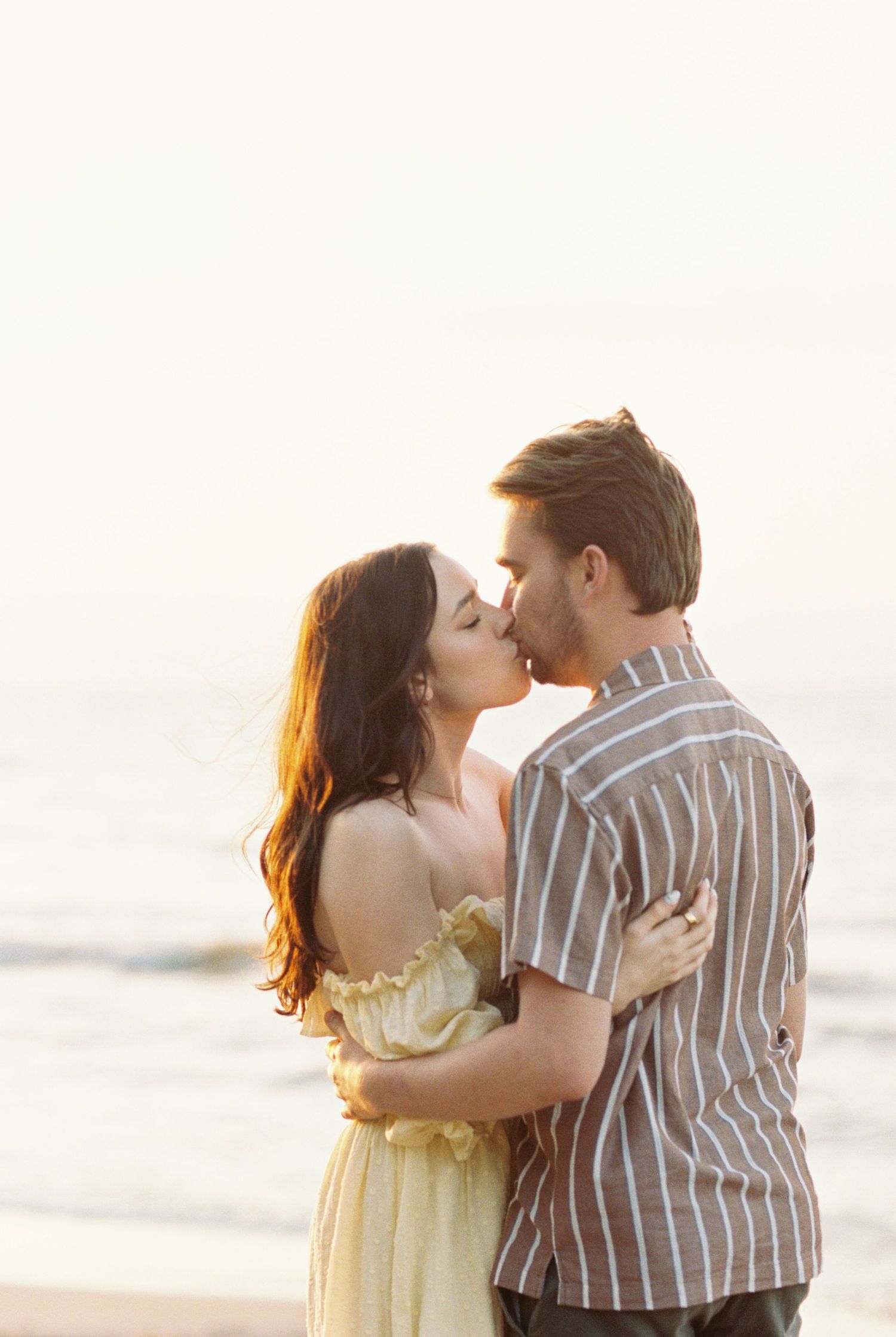 A couple shares a romantic kiss on the beach at sunset while wearing a yellow dress and striped shirt.
