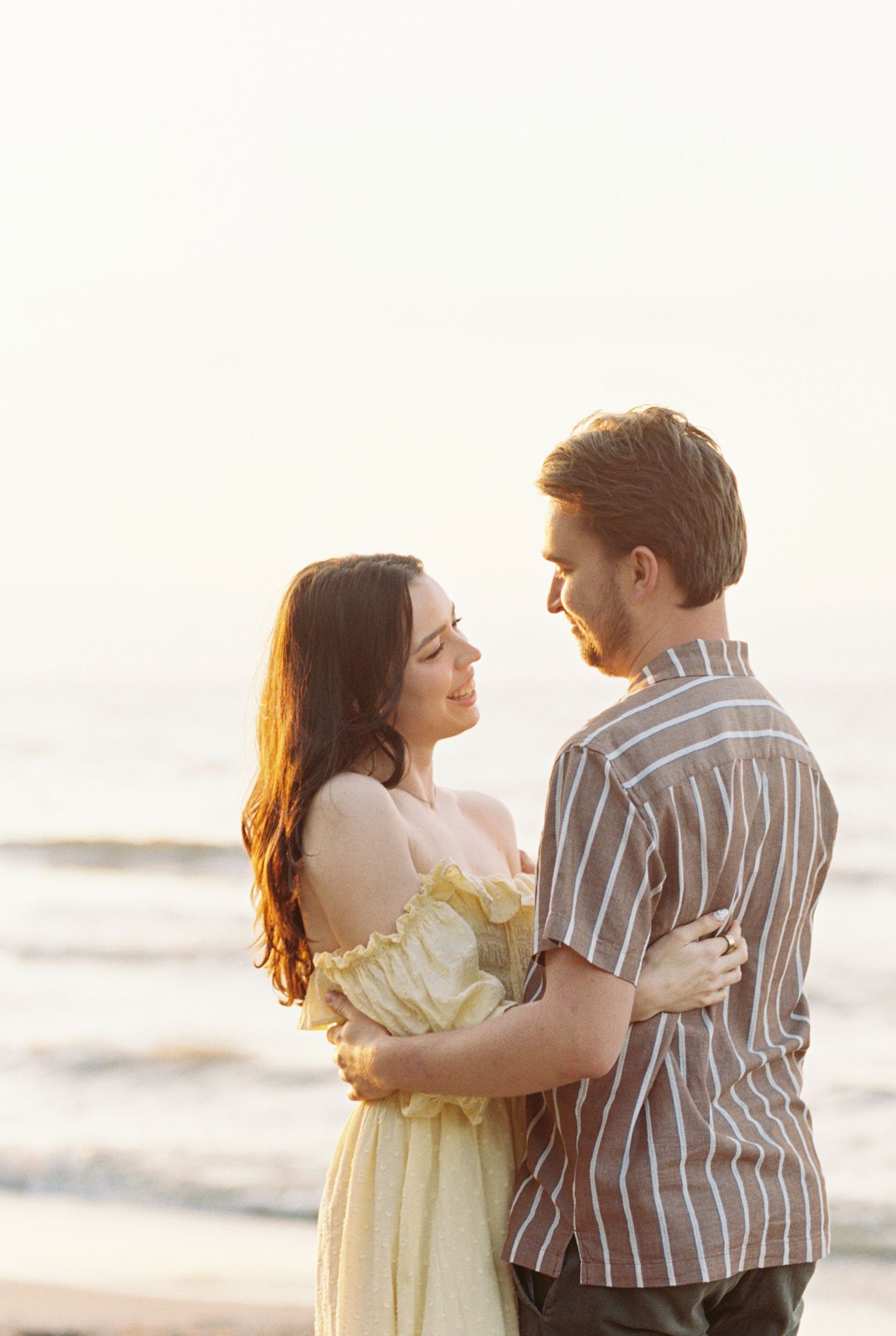 A couple embraces on the beach at sunset, with warm golden light illuminating them as they share an intimate moment.