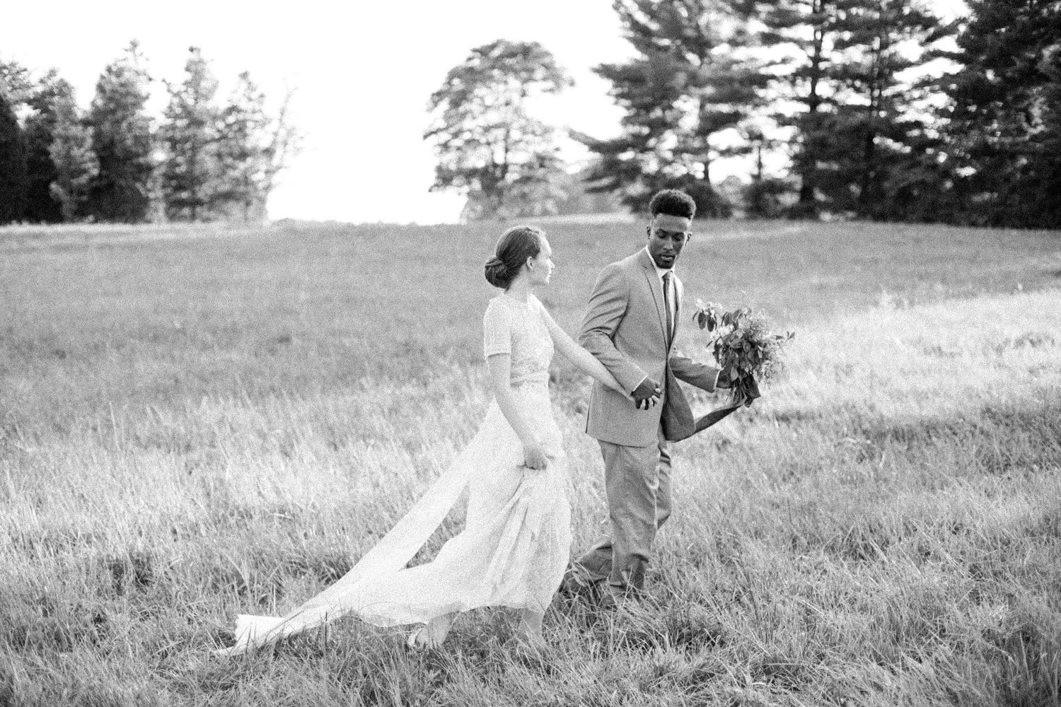 A black and white photograph of a couple walking through a sunlit meadow with pine trees in the background. Kentucky Wedding Photographer
