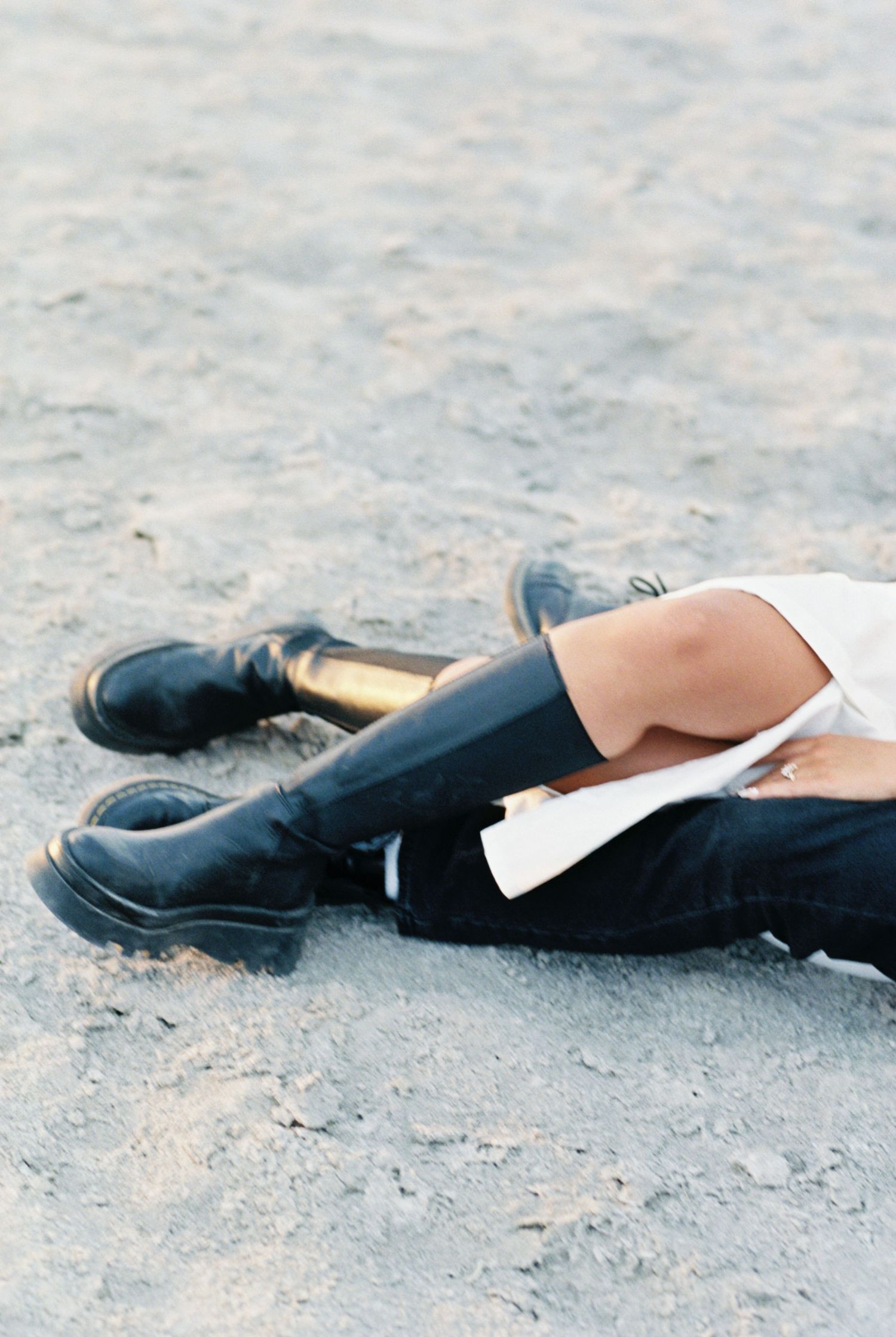 Person wearing black boots and pants resting on a sandy gray surface.