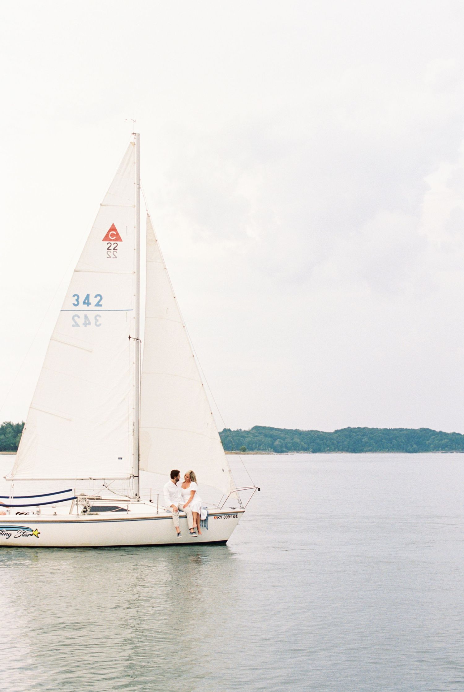 A KY sailboat with white sails glides across calm waters with distant shoreline visible. Kirstie Whitaker Photography