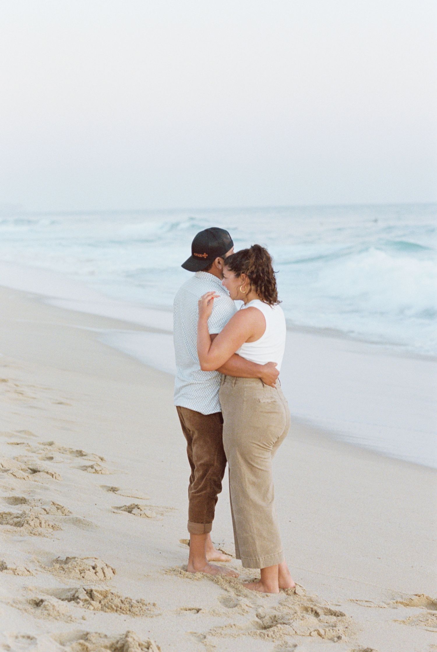 Couple embracing on sandy beach at sunset with ocean waves in background. Kirstie Whitaker Photography