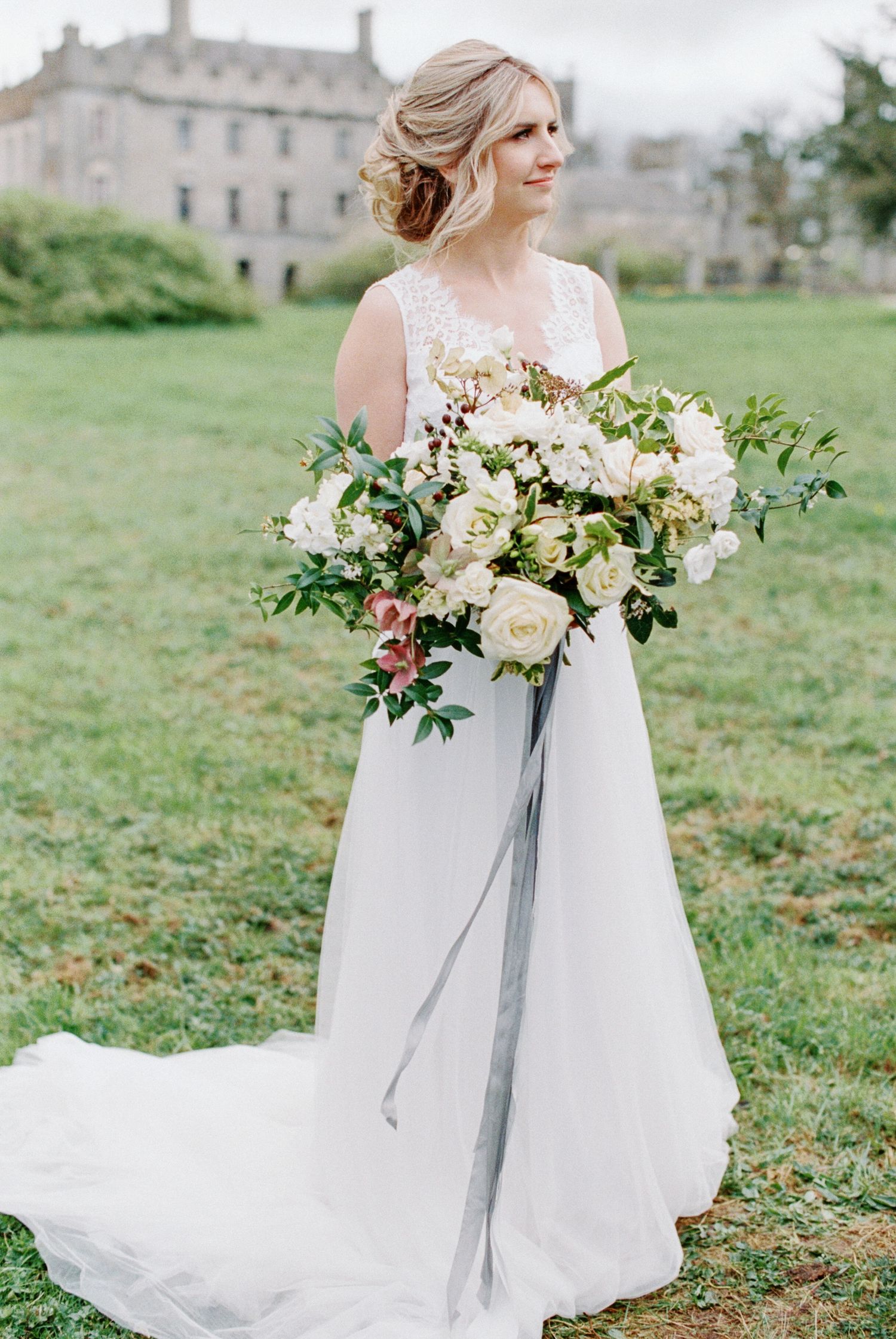 Elegant bride in white lace gown holds luxurious bouquet of cream roses and greenery on castle grounds.