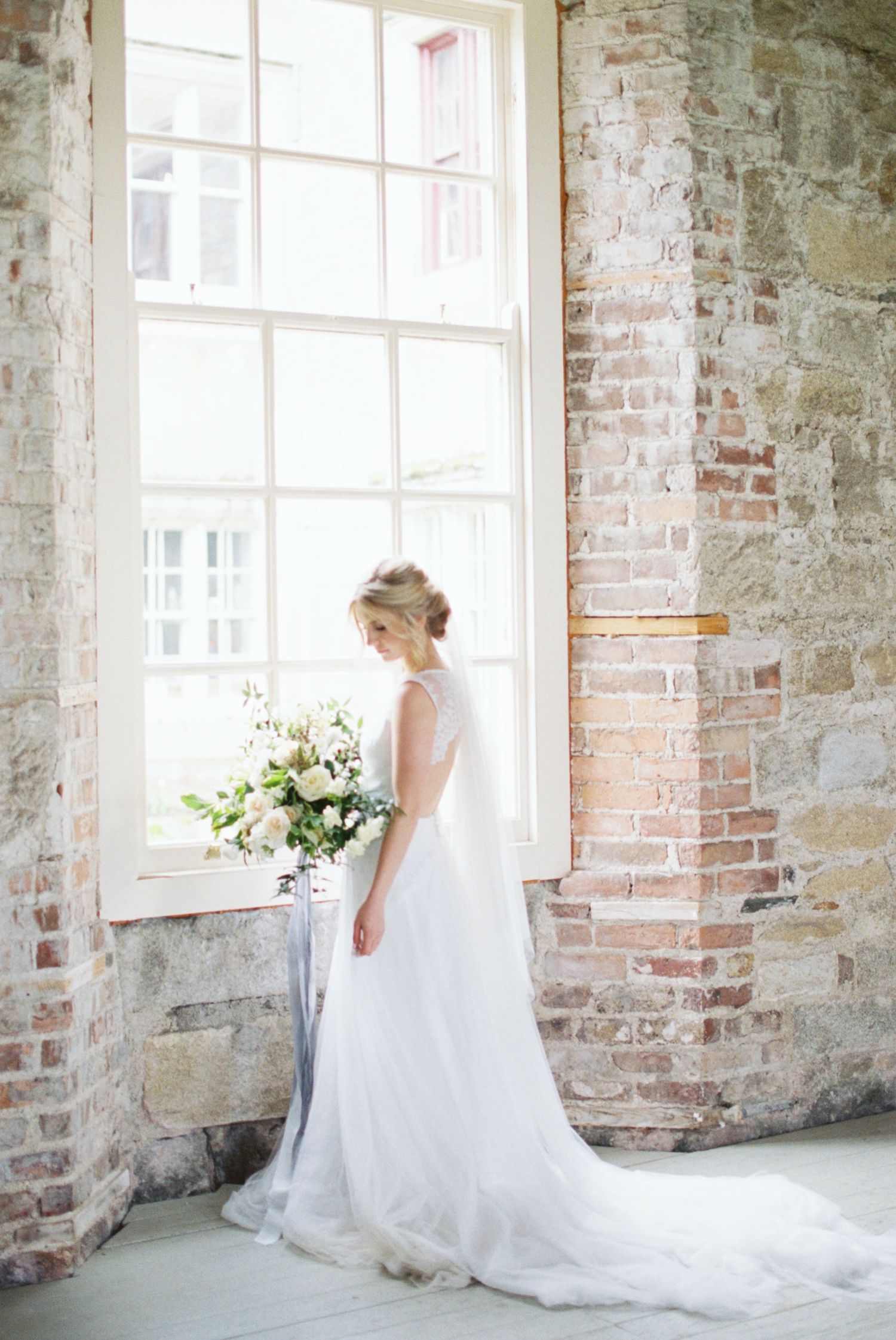Elegant white wedding gown with flowing train photographed against exposed brick wall and bright window in rustic setting.