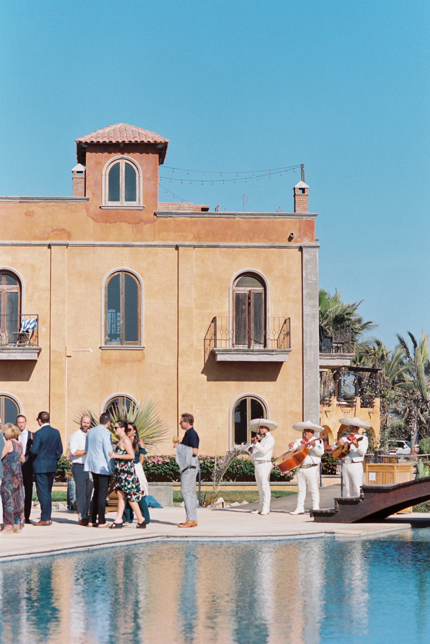 Guests gather around a Mediterranean-style villa with a swimming pool during an outdoor event on a sunny day.