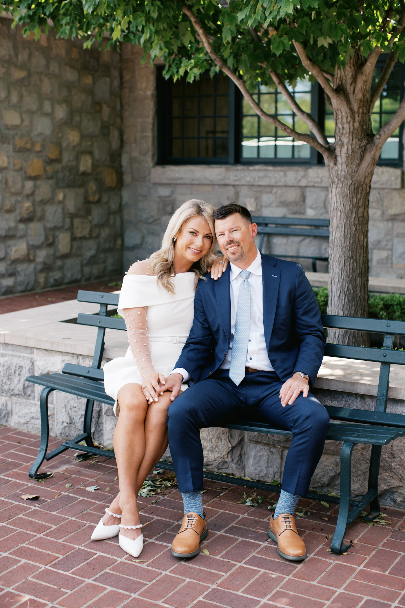 A couple sits together on a green park bench against a stone wall and tree on a brick patio.