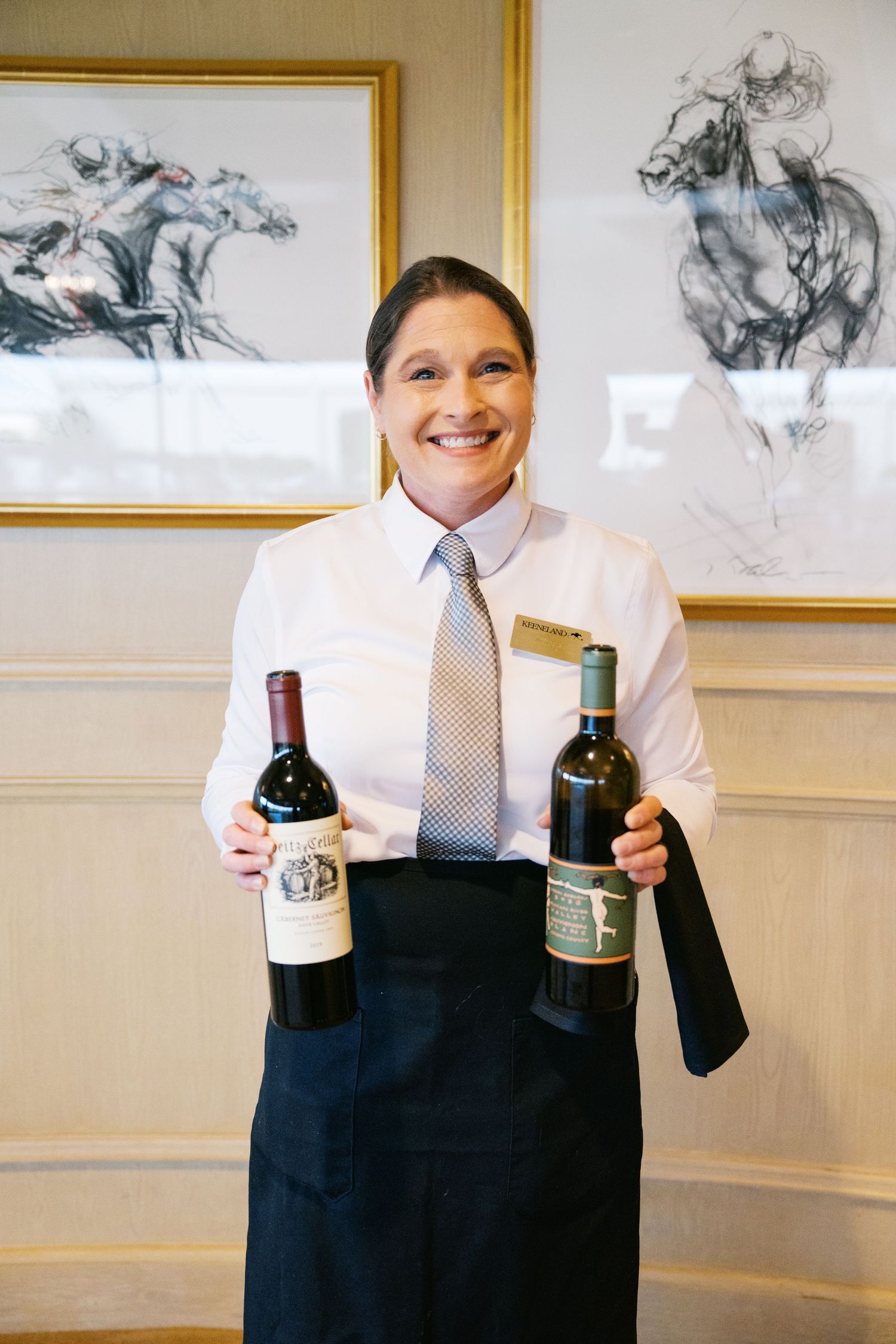 A restaurant server in professional attire displays two bottles of wine in front of equestrian artwork on the wall.