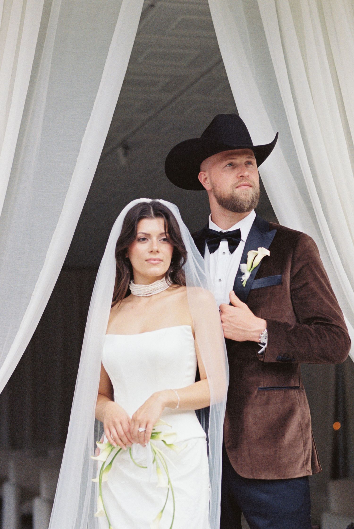 A couple in wedding attire poses under white curtains, with the bride in white and groom wearing a brown suit.