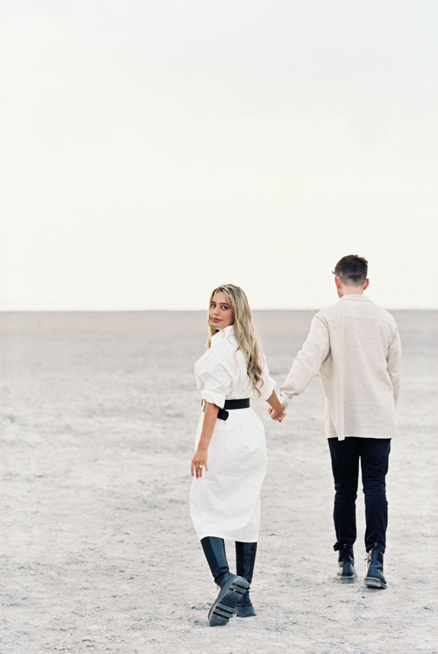 A couple walking hand in hand across a desert landscape wearing coordinated white outfits. Kirstie Whitaker Photography