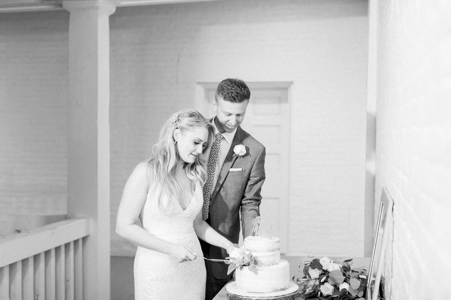 A couple in wedding attire cuts their white wedding cake together on a porch in black and white photography. Nashville Wedding Photographer