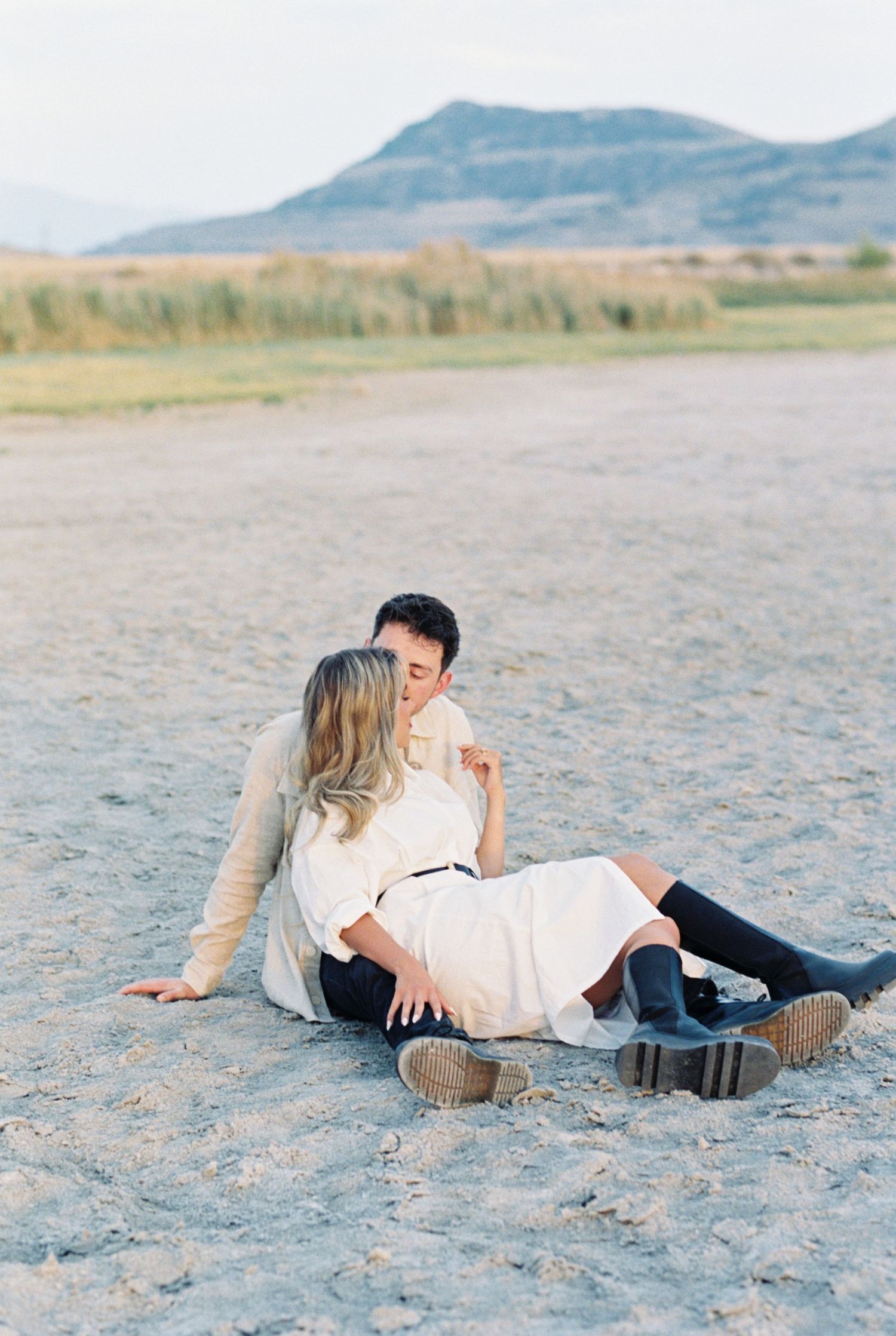 A romantic couple sits together on sandy ground with mountains in the distant background.