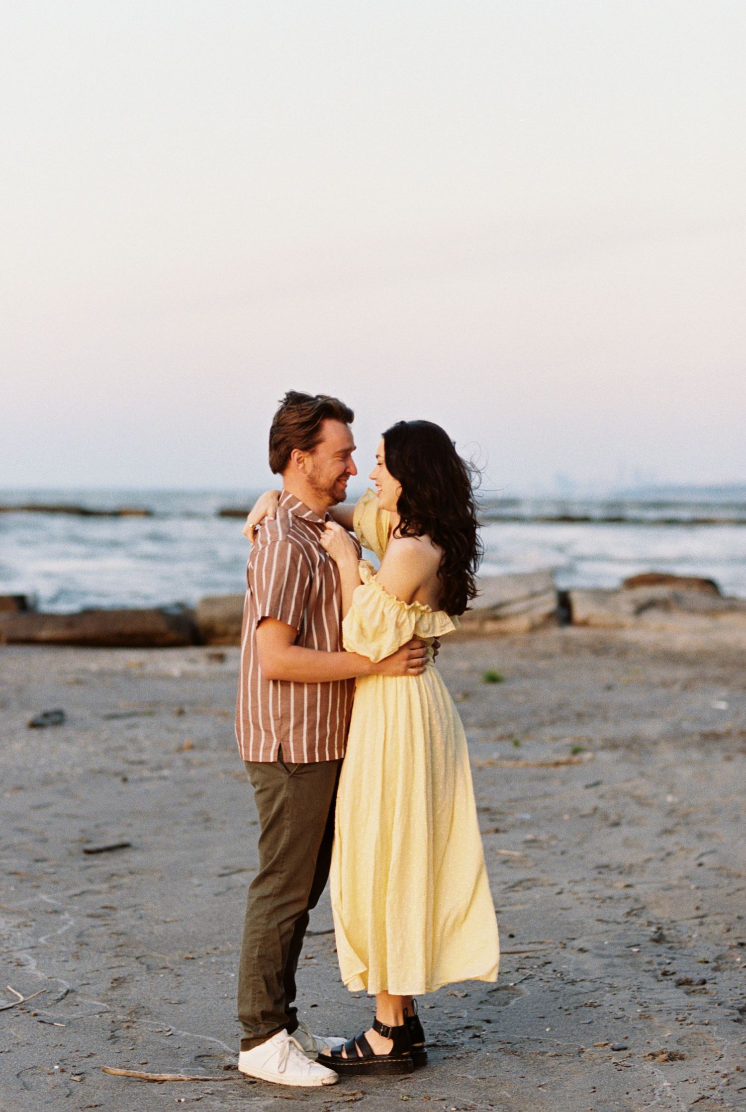 A couple embraces on a rocky beach at sunset, with the woman wearing a flowing yellow dress and man in stripes.