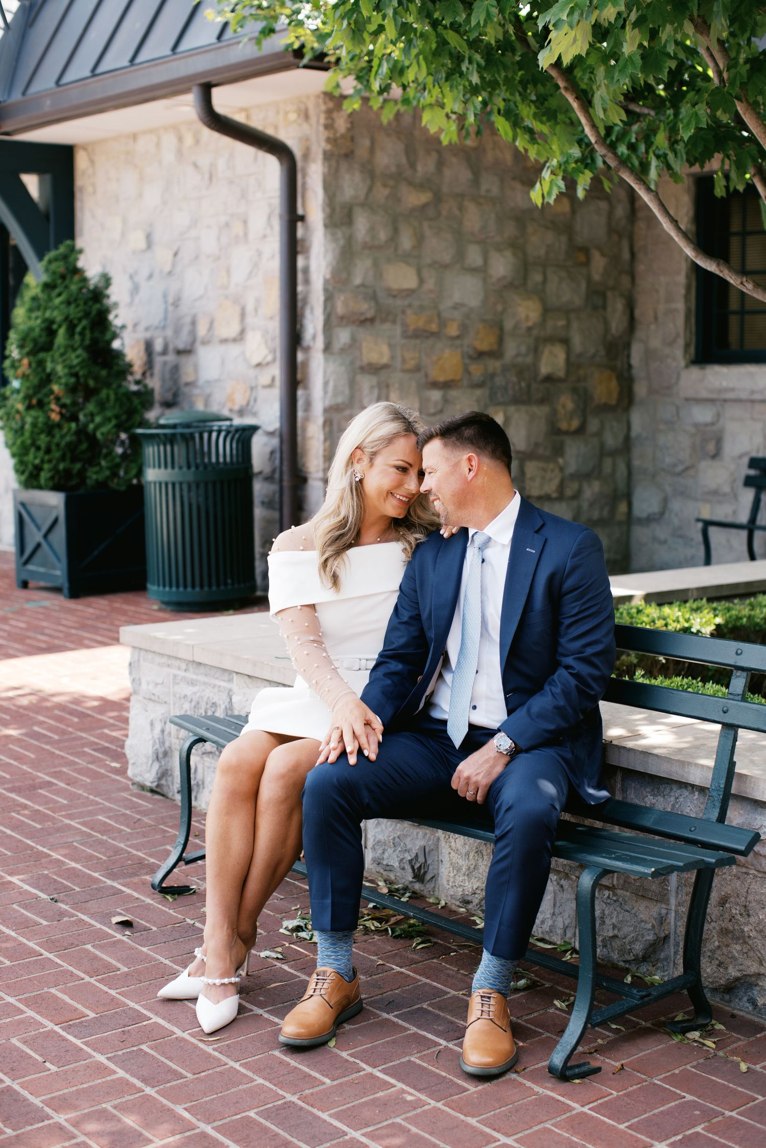 A couple sits together on a bench near a stone wall and brick walkway in a romantic outdoor setting.