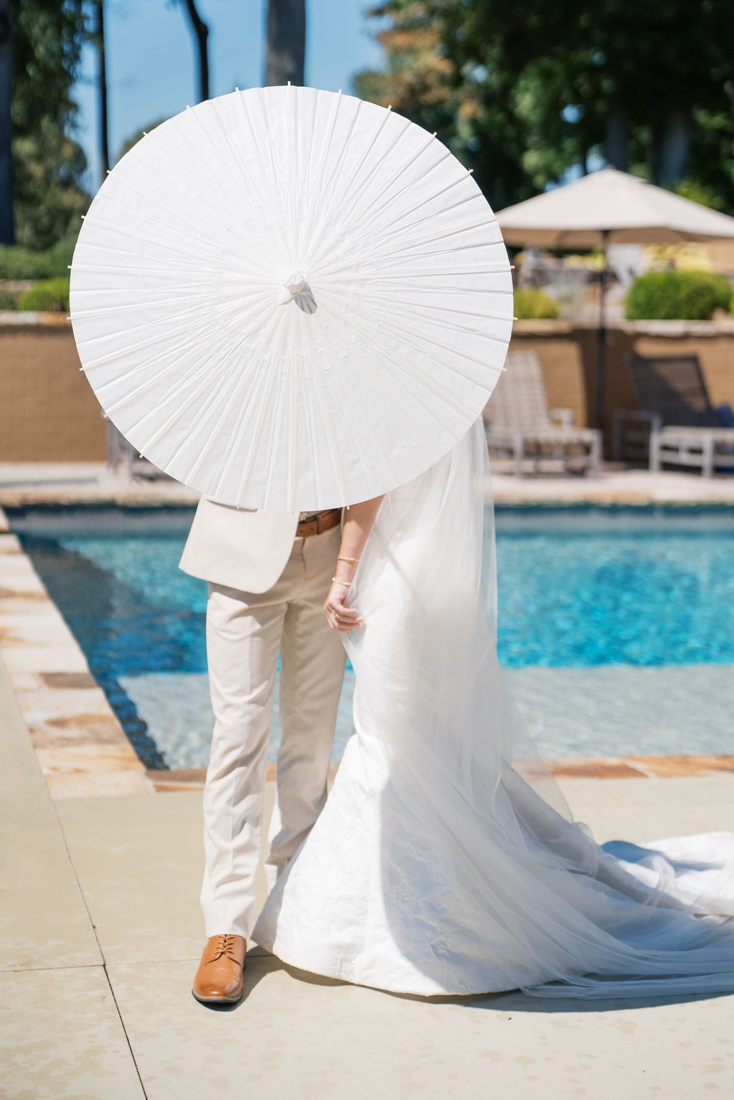 A couple shares an intimate moment behind a white paper parasol by a swimming pool on their wedding day.