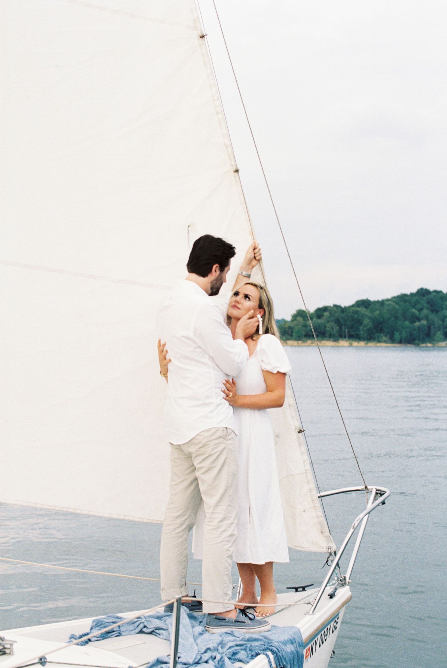 A romantic moment aboard a sailboat with two people in white outfits standing against a lake backdrop.