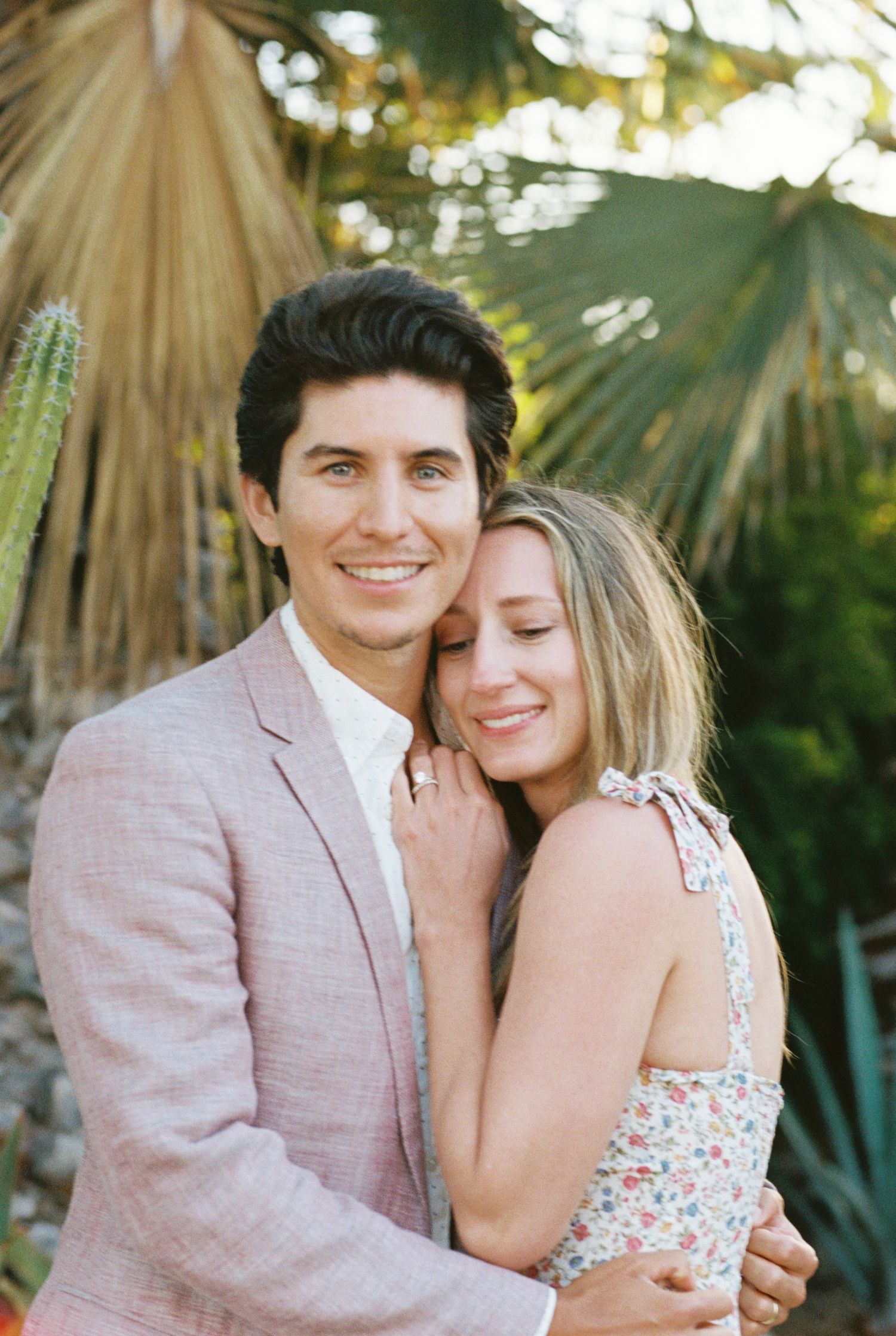 A romantic couple embraces outdoors at sunset, with the woman in a floral dress and man wearing a light pink blazer.
