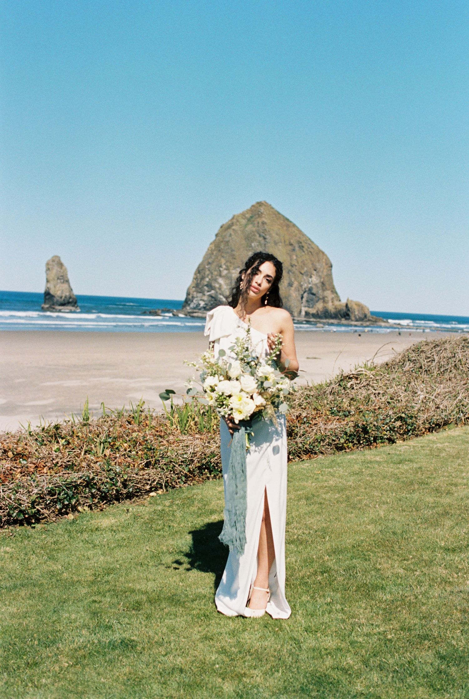 A person in a white dress holds a bouquet of flowers on a beach with Haystack Rock visible in the background at Cannon Beach.