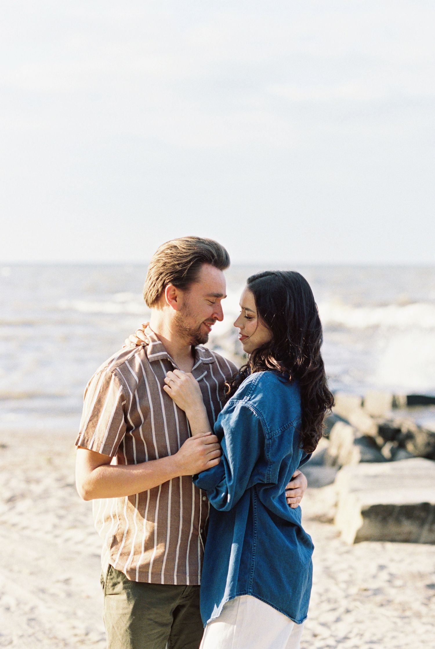Romantic couple embraces on a sunny beach with waves and rocks in the background during golden hour.