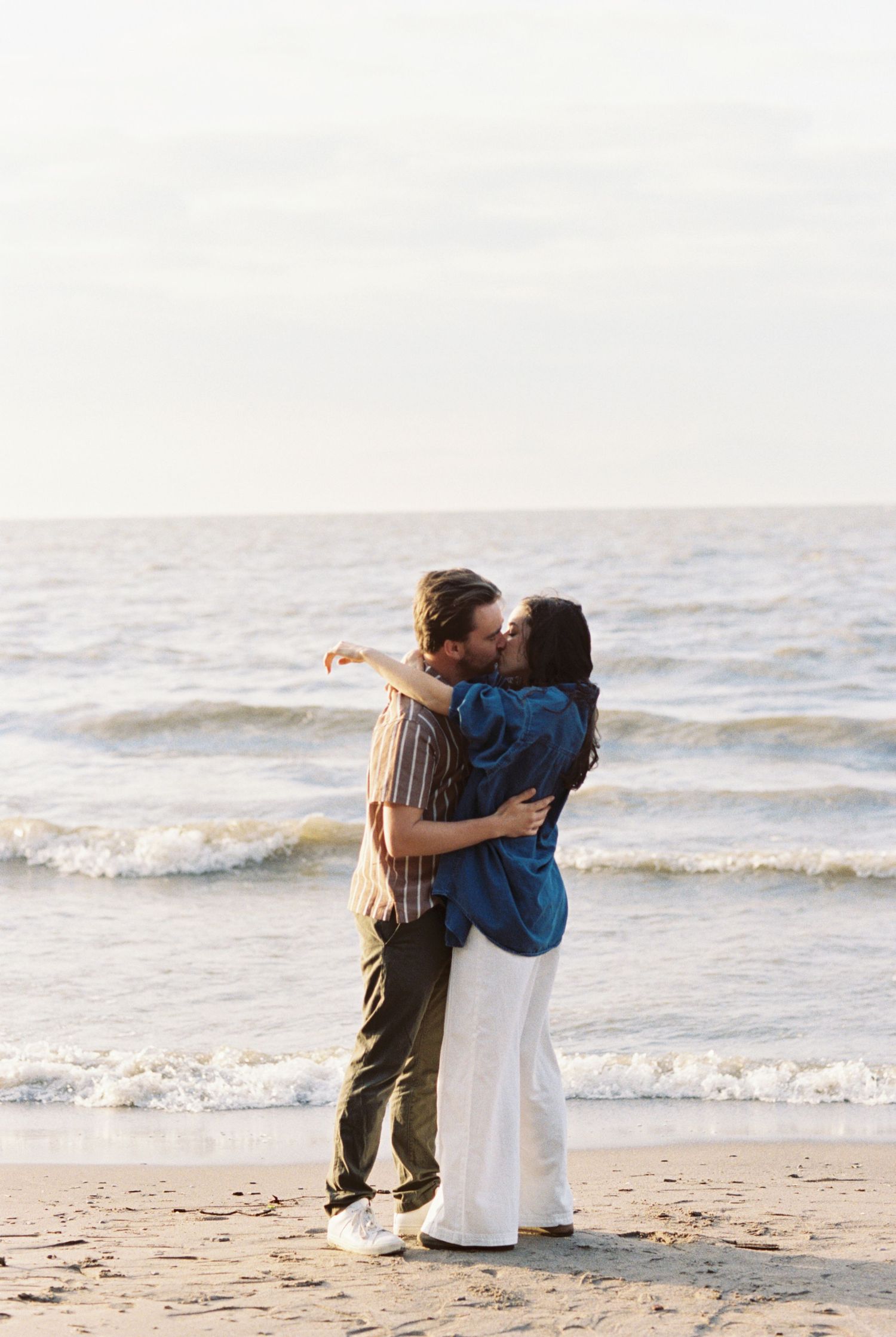A romantic couple shares an intimate embrace on a sandy beach at sunset with gentle ocean waves in the background.