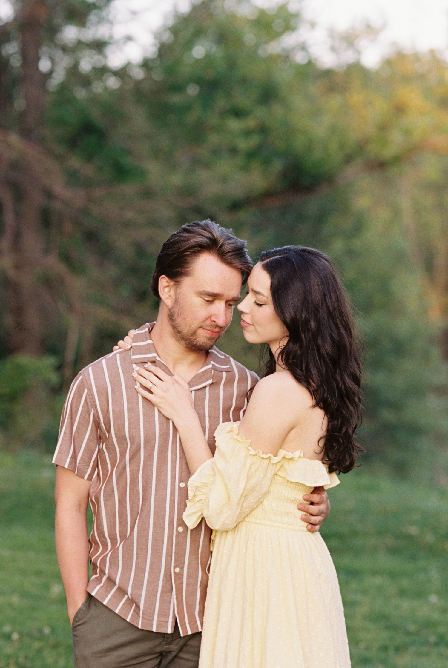 A romantic couple standing close together outdoors, with the woman wearing a yellow dress and the man in a brown striped shirt.