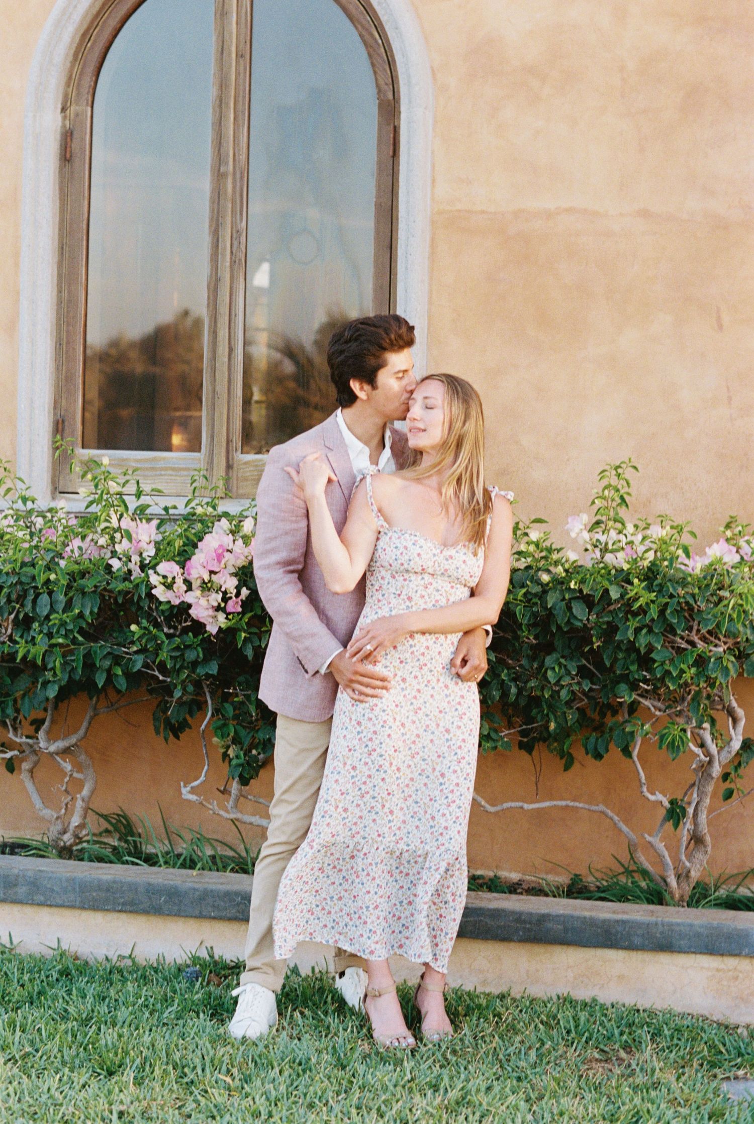 A romantic moment between a couple standing by an arched window with pink flowers and greenery against a peach-colored wall.