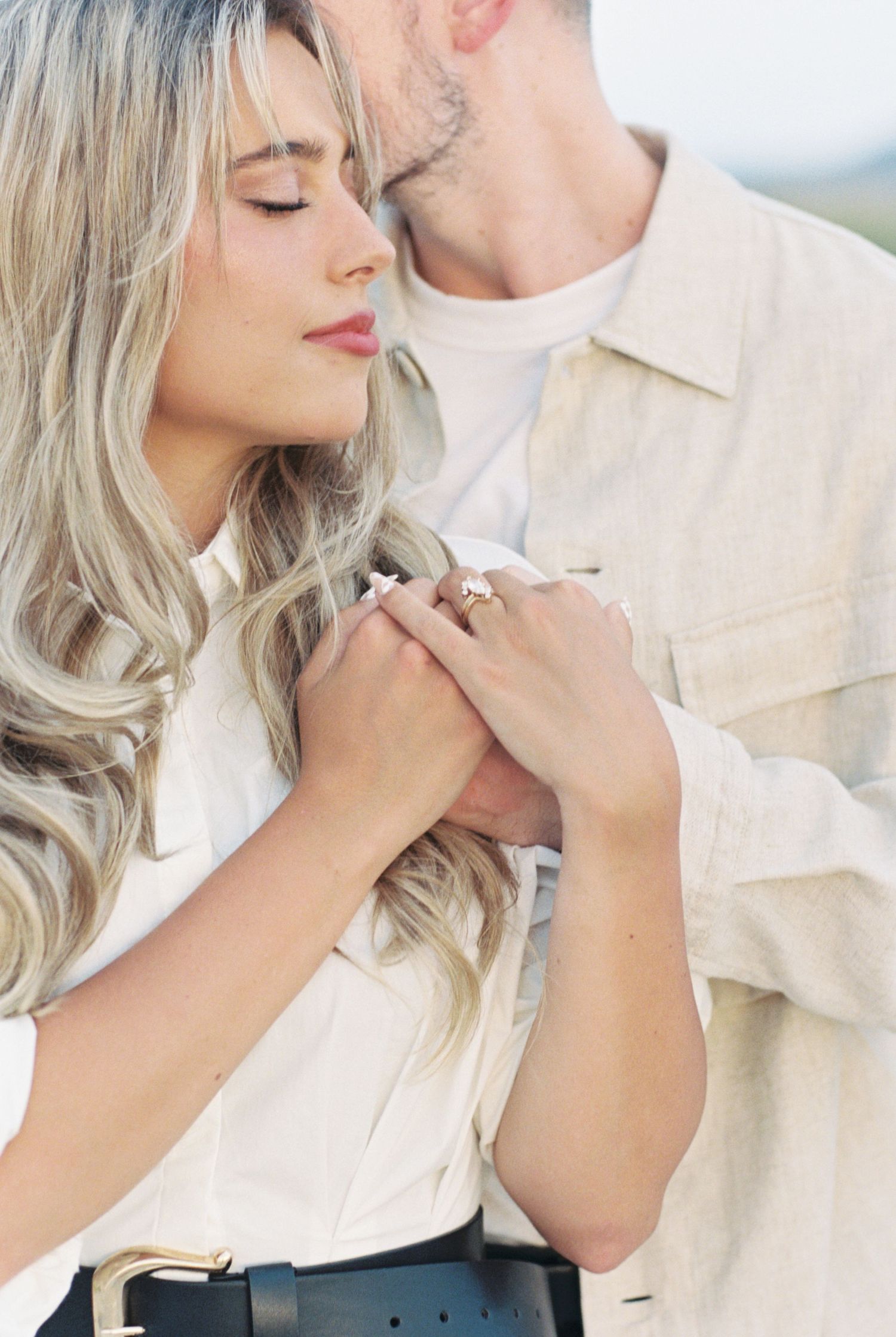 A tender romantic moment captured in soft lighting, with flowing blonde hair and light neutral-toned clothing.